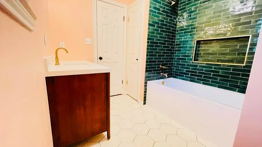 Bathroom with a white bathtub, green tiled wall, white hexagon floor tiles, a wooden vanity with a white top and gold faucet, and two white doors.