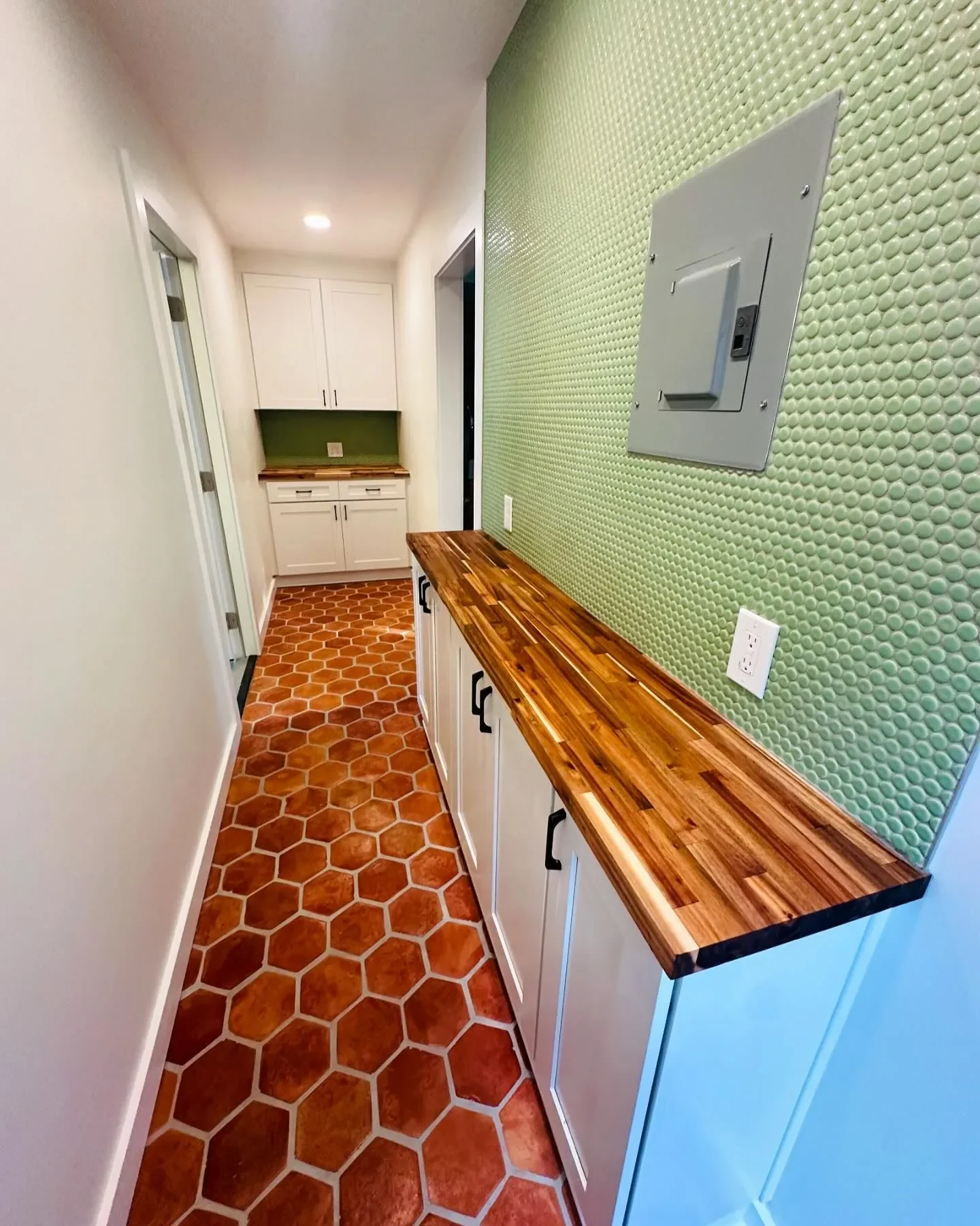 Narrow kitchen hallway with hexagonal terracotta tile flooring, white cabinets, green textured wall, wooden countertop, and two doors at the end.