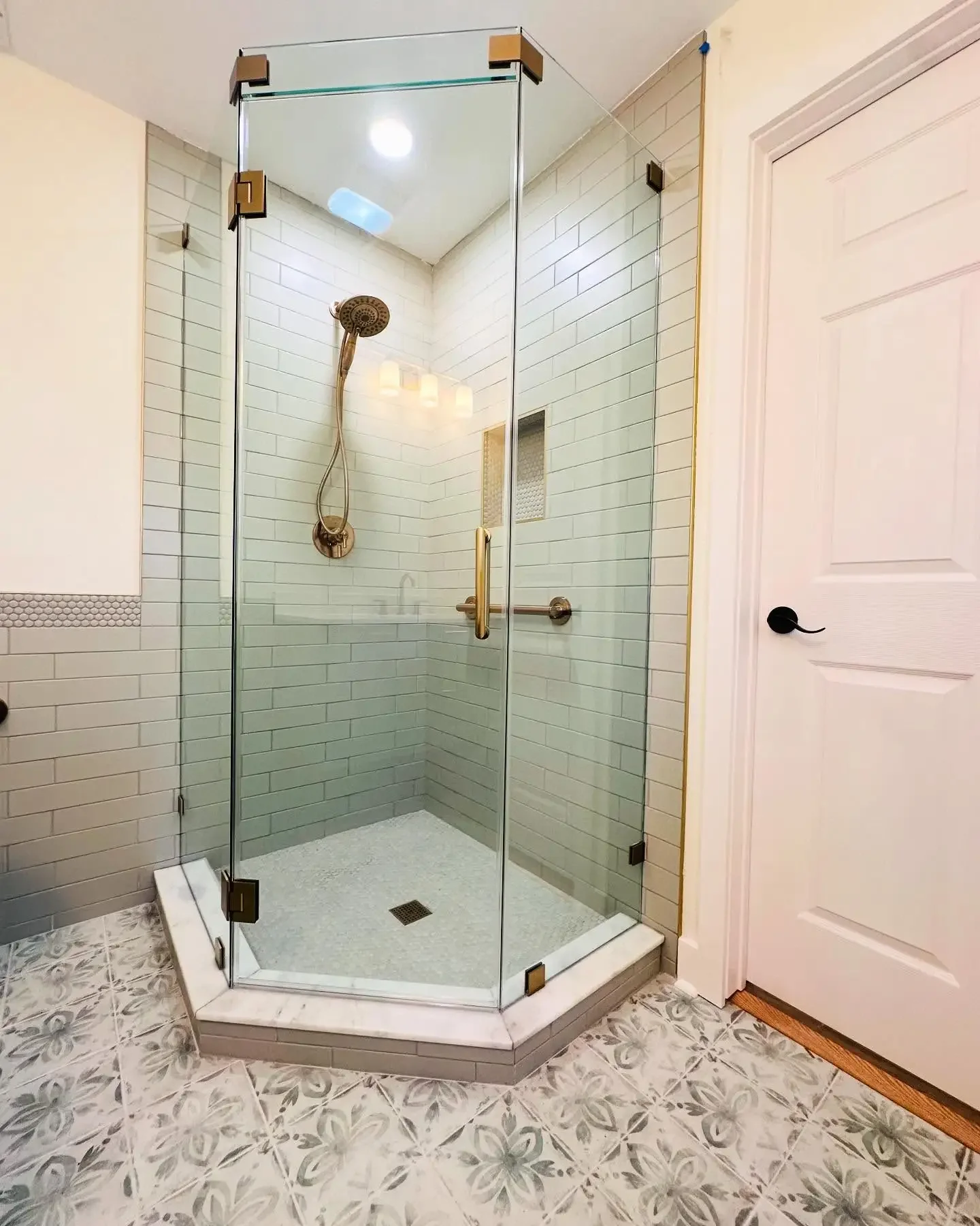 A corner shower with glass doors, brass hardware, tiled walls, a wall-mounted showerhead, a niche, and a grab bar, inside a bathroom with patterned tile flooring and a white door.