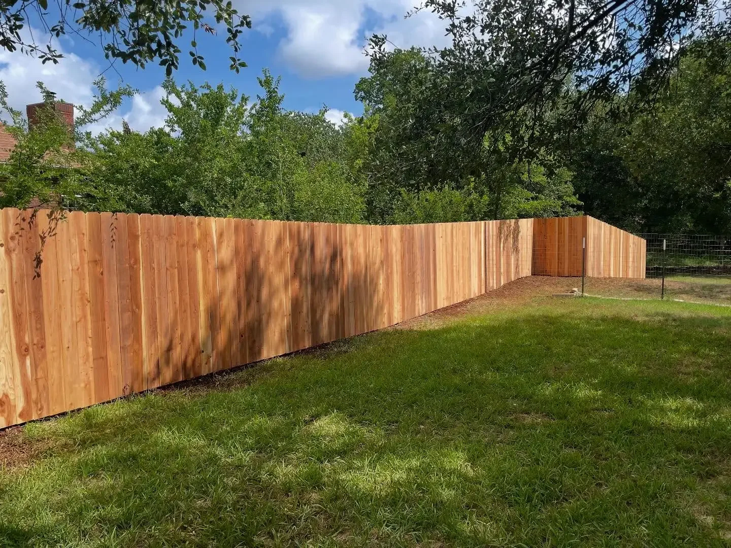 Wooden privacy fence in a backyard with green grass and trees, under a partly cloudy sky.
