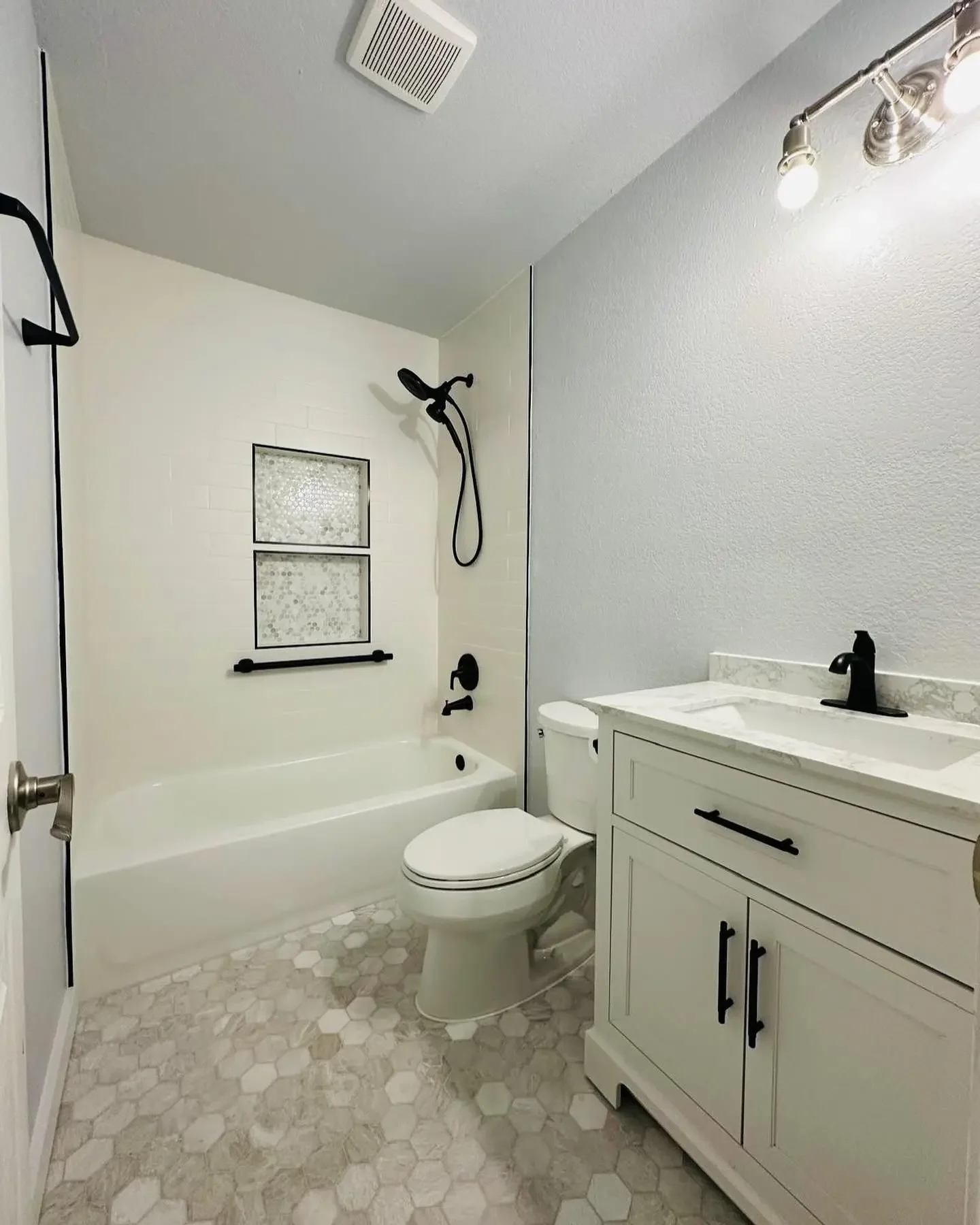 A modern bathroom with a bathtub and shower combo, a toilet, and a white sink vanity with black fixtures. The floor has hexagonal tiles, and there's a wall-mounted light above the mirror.