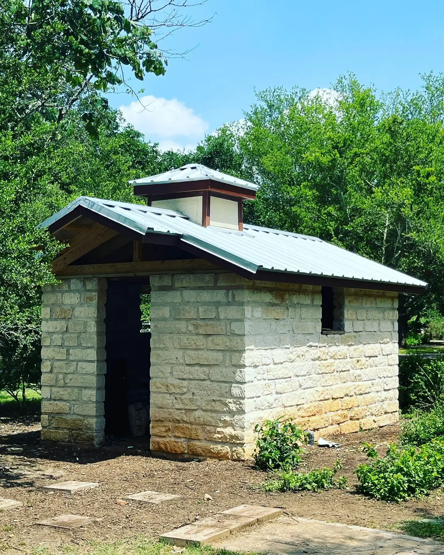 A small stone building with a metal roof and a small vent on top, surrounded by green trees and shrubs, under a blue sky.