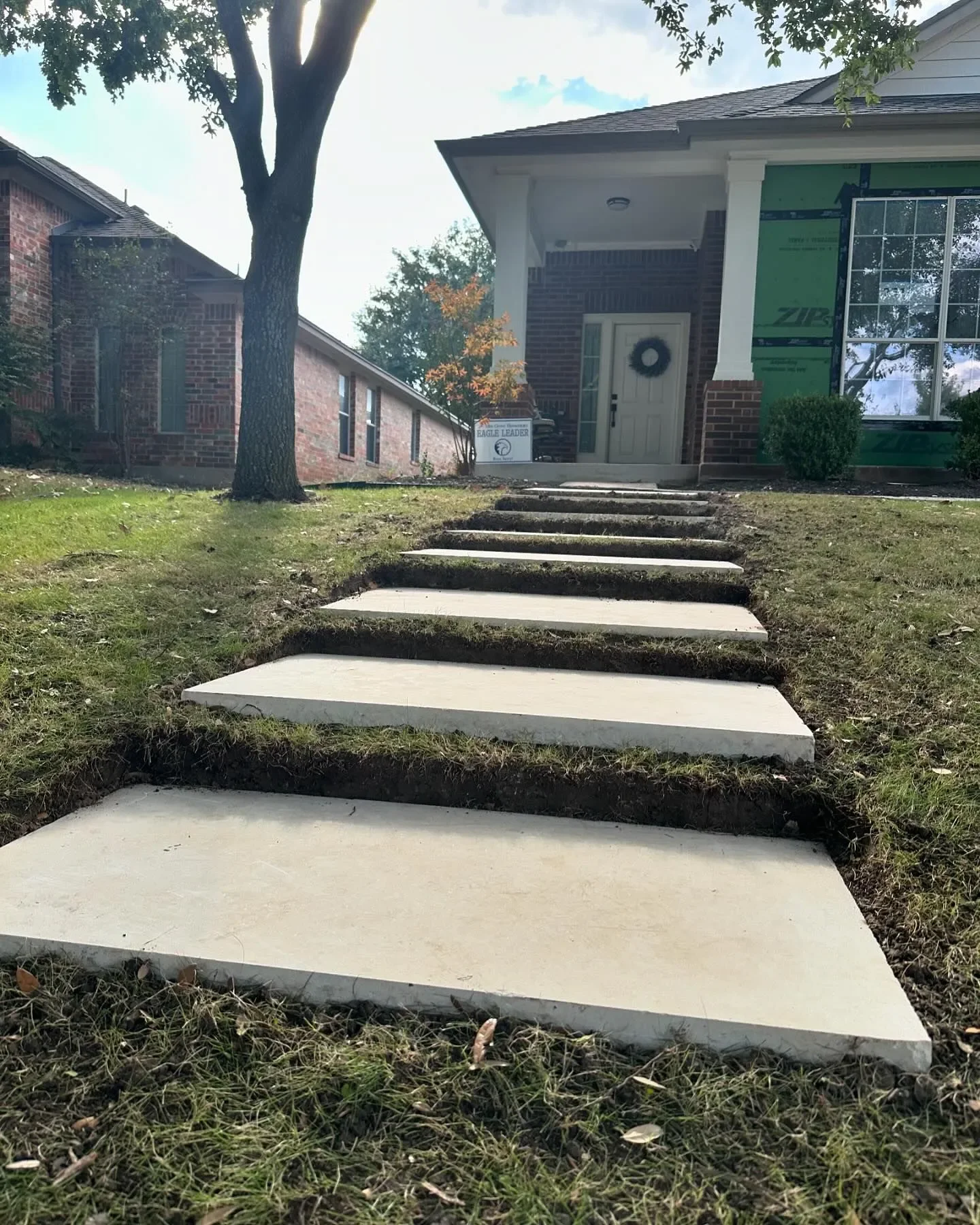 Concrete stepping stones leading up to the front porch of a house with a wreath on the door, a tree on the lawn, and a partially constructed section of the house with green exterior sheathing.