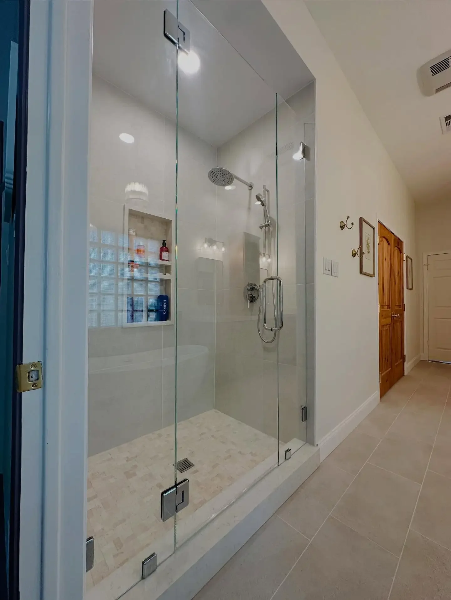 Modern walk-in shower with glass doors, rain showerhead, built-in shelf with toiletries, and beige tile flooring, in a home bathroom.