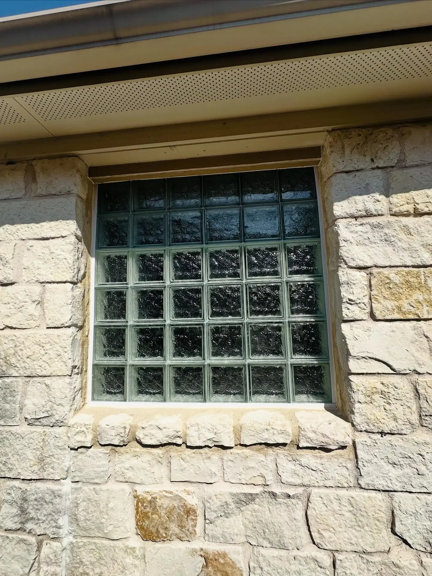 A window made of glass blocks set into a stone wall with a beige stone frame, reflecting trees outside.