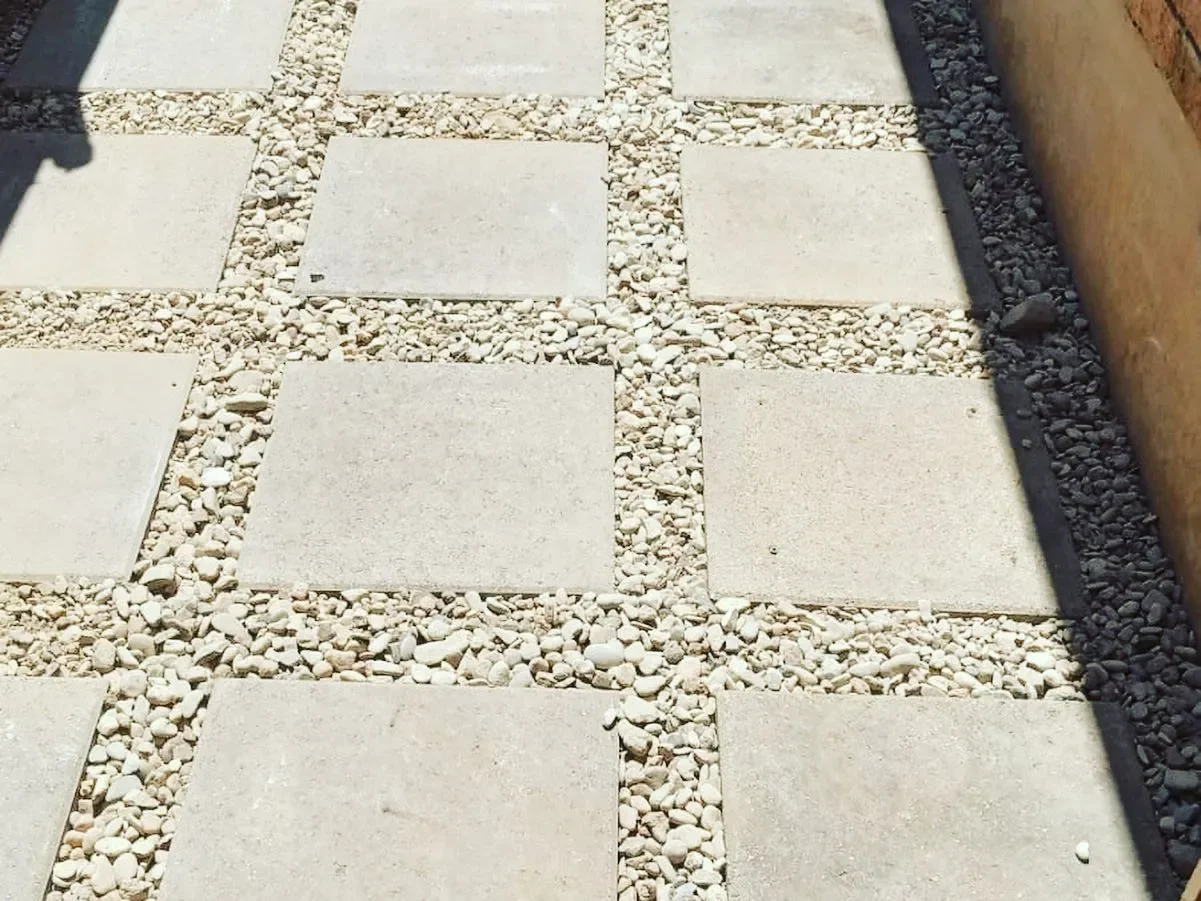 A pathway with large square concrete pavers separated by gravel filled gaps, bordered on the right by a brick wall and on the left by a shadow.