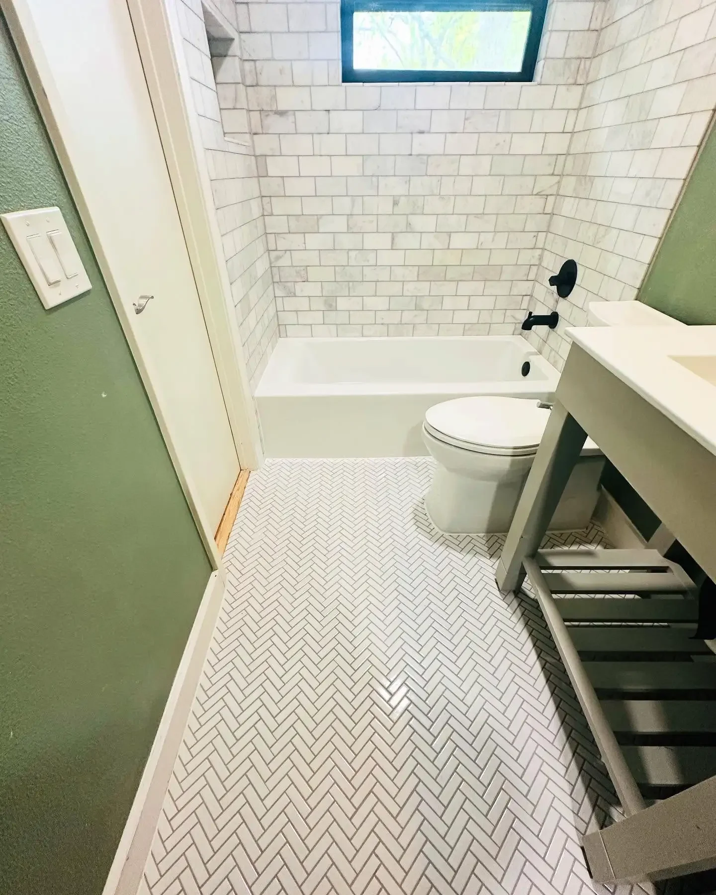 Bathroom with green walls, white herringbone tile floor, bathtub with white subway tile surround, black fixtures, a small gray vanity, and a small window.