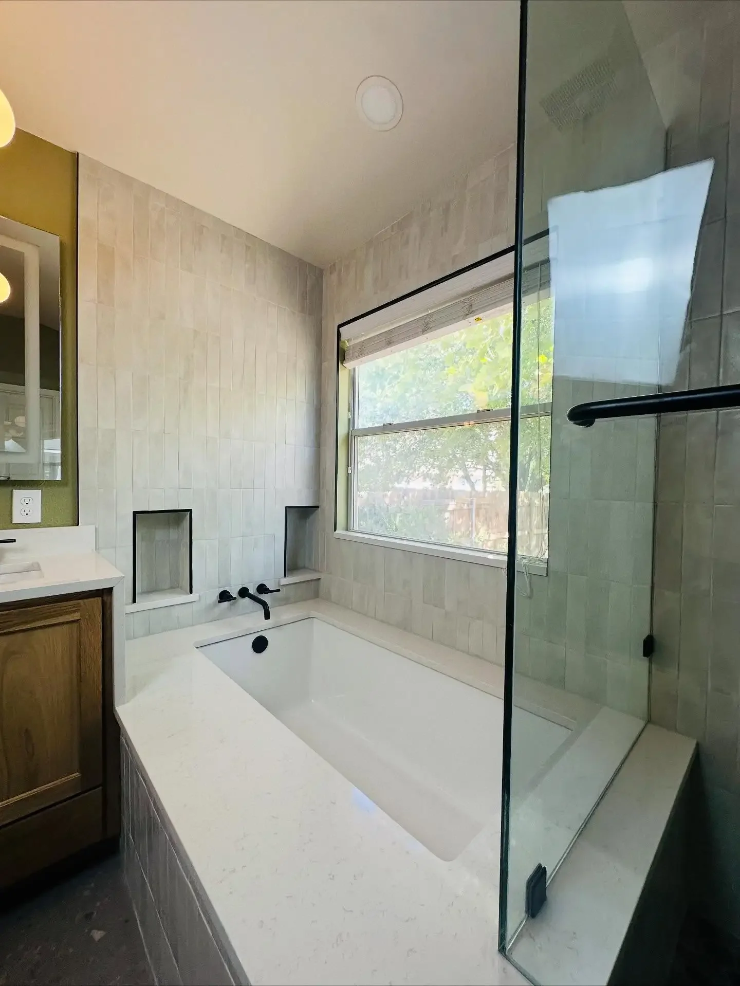 A modern bathroom featuring a bathtub with a black faucet, a large window with greenery outside, and a glass-enclosed shower stall.