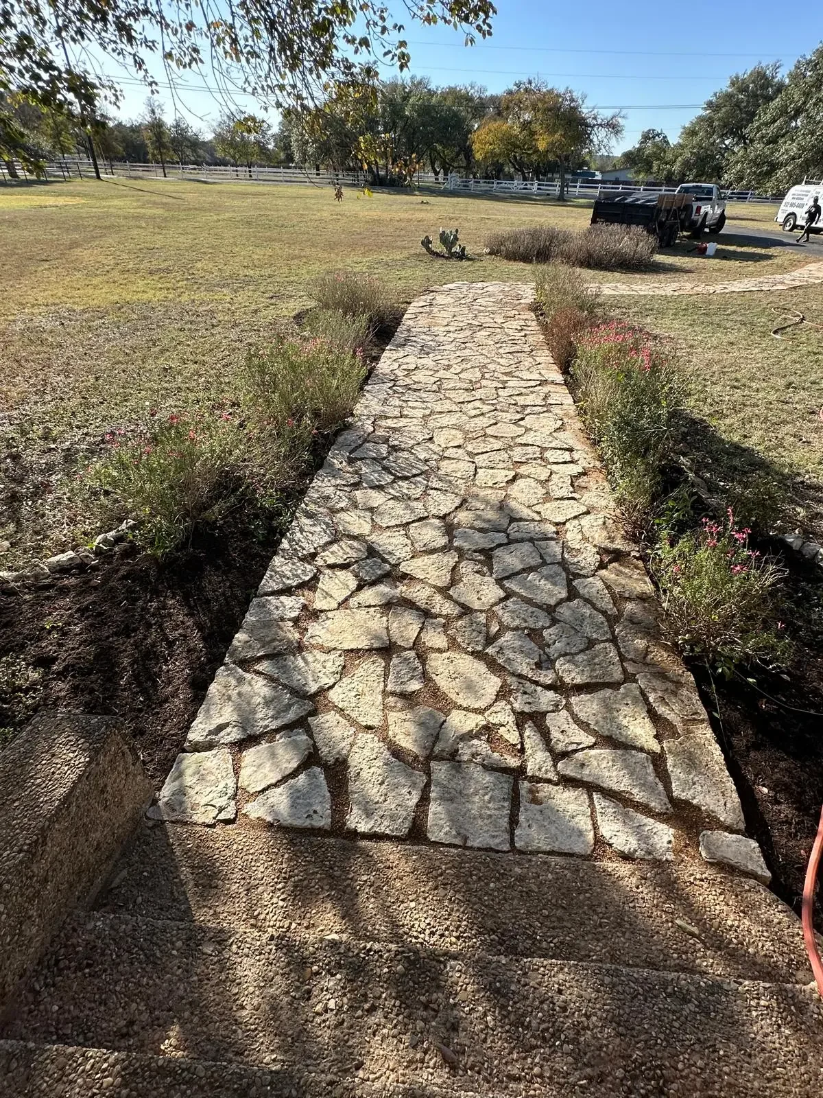 Stone pathway leading through landscaped area with flowering plants on sides, extending towards a grassy yard with trees and parked vehicles in the background.