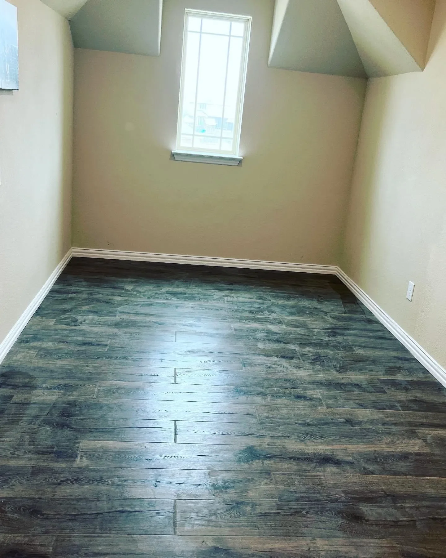 Empty small room with beige walls, a window on the back wall, dark wood flooring, white baseboards, and an electrical outlet on the right wall.