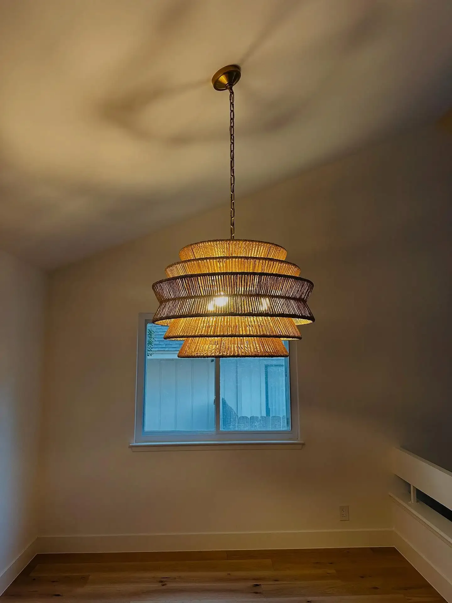 A hanging pendant light fixture with multiple layered shade panels, illuminated and casting a glow on an empty room with a window and wooden floor.
