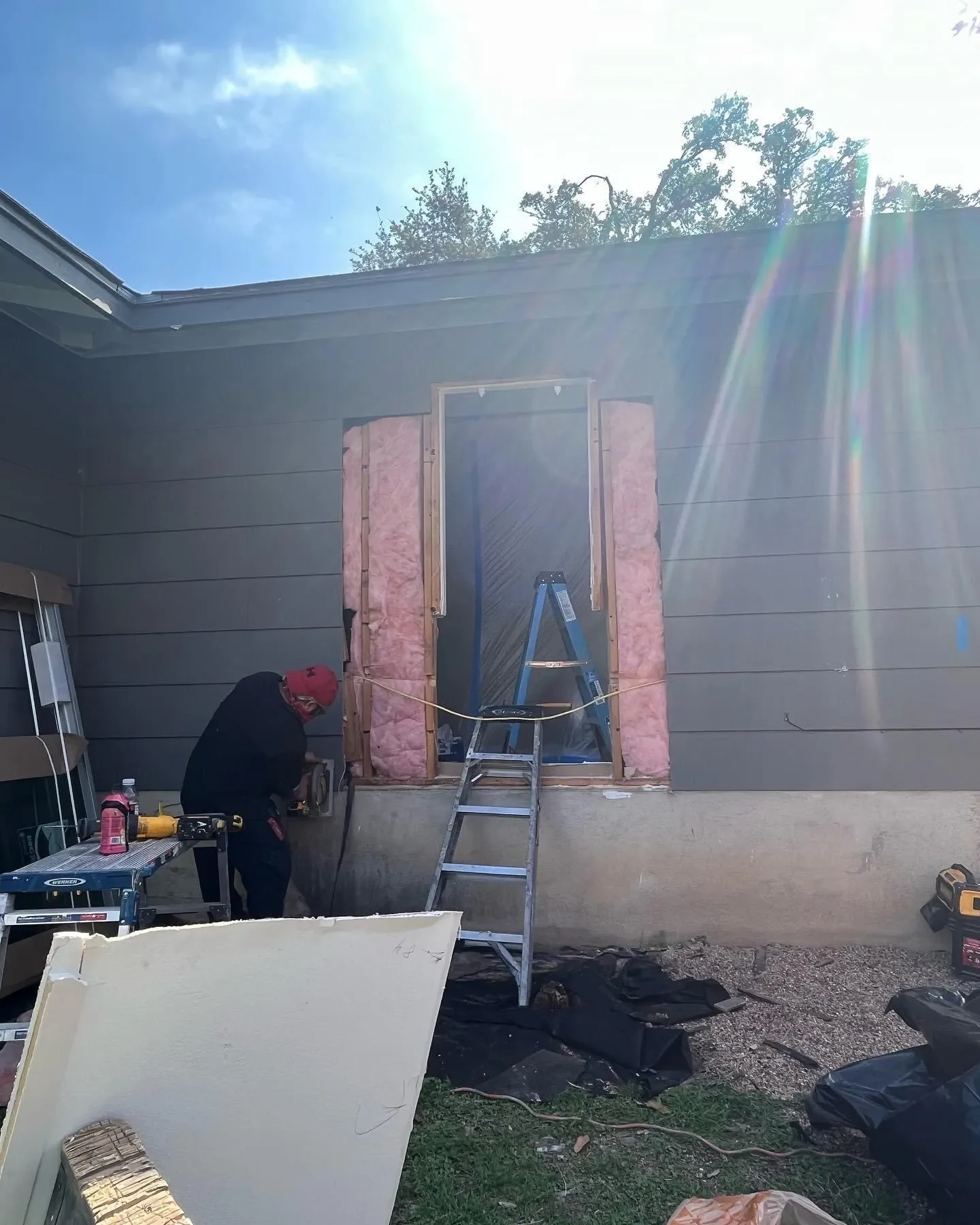 A person working on the exterior wall of a house, installing a new window or door frame with pink insulation around the opening, using tools, with a ladder set up in front, and construction materials and equipment around.