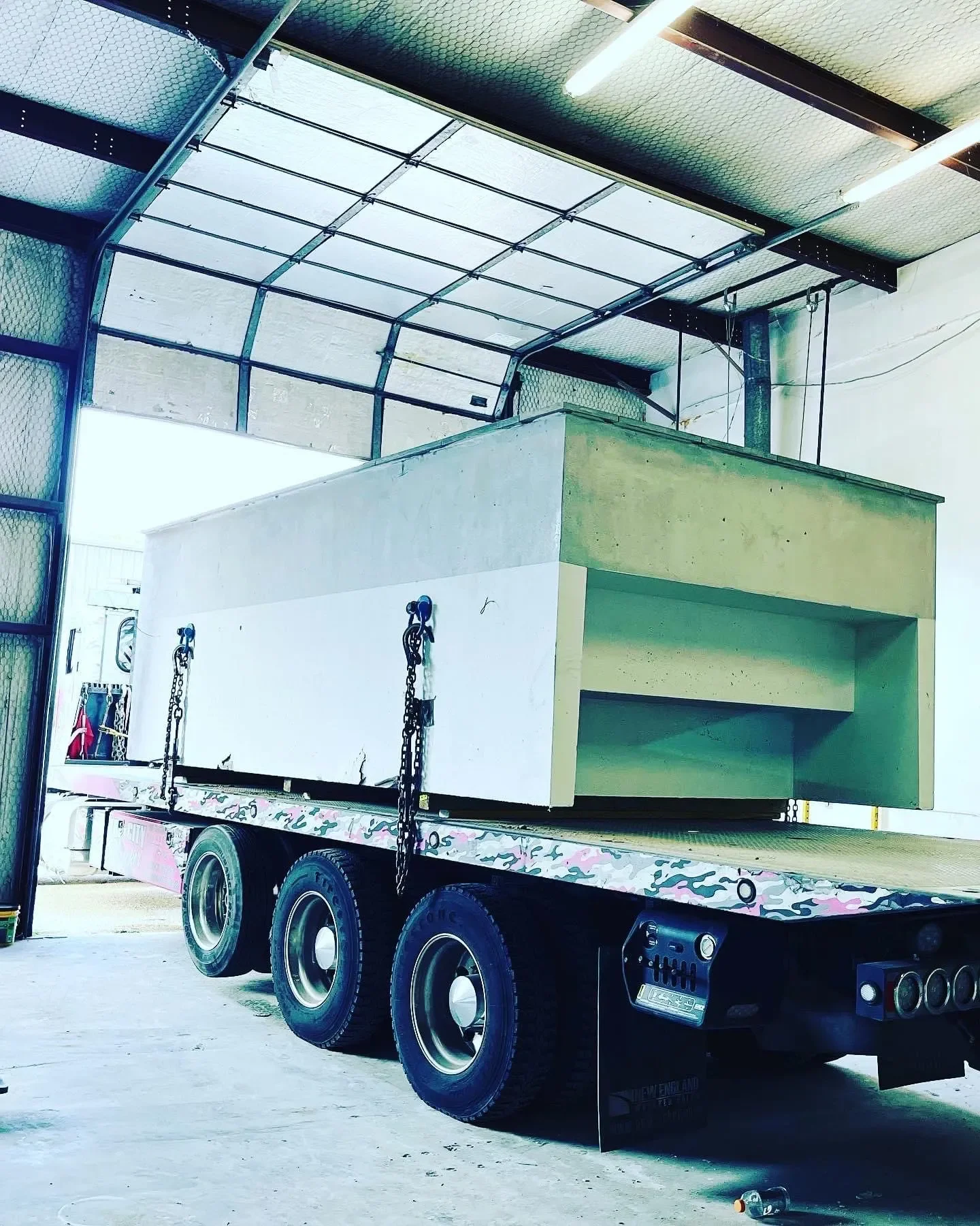 A flatbed truck inside a warehouse carrying a large concrete structure with a rectangular cutout, attached with chains.