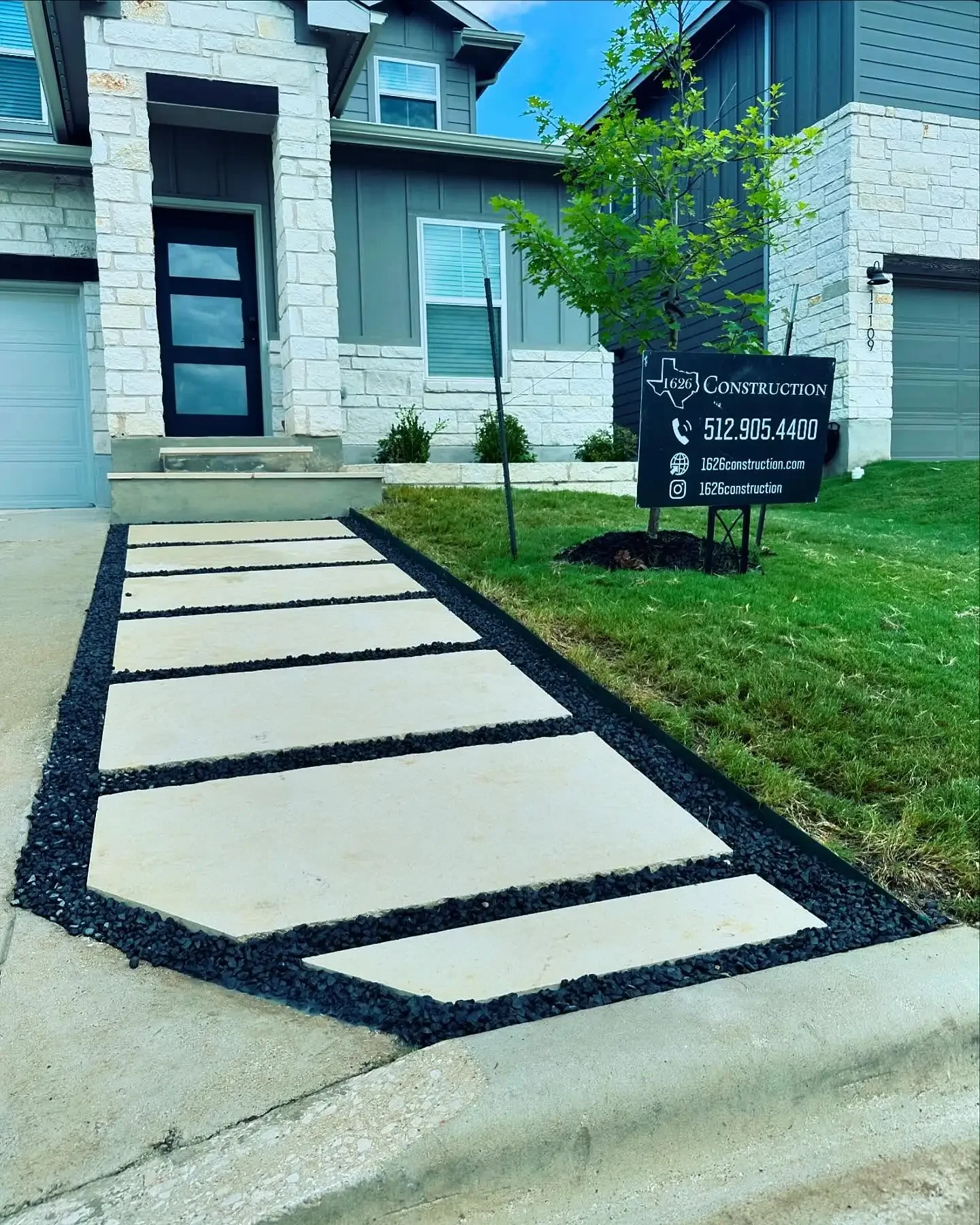 Concrete pathway with evenly spaced stepping stones leading to the front door of a modern house with stone and siding exterior. There is a grass lawn and a small tree with a construction sign in the yard.