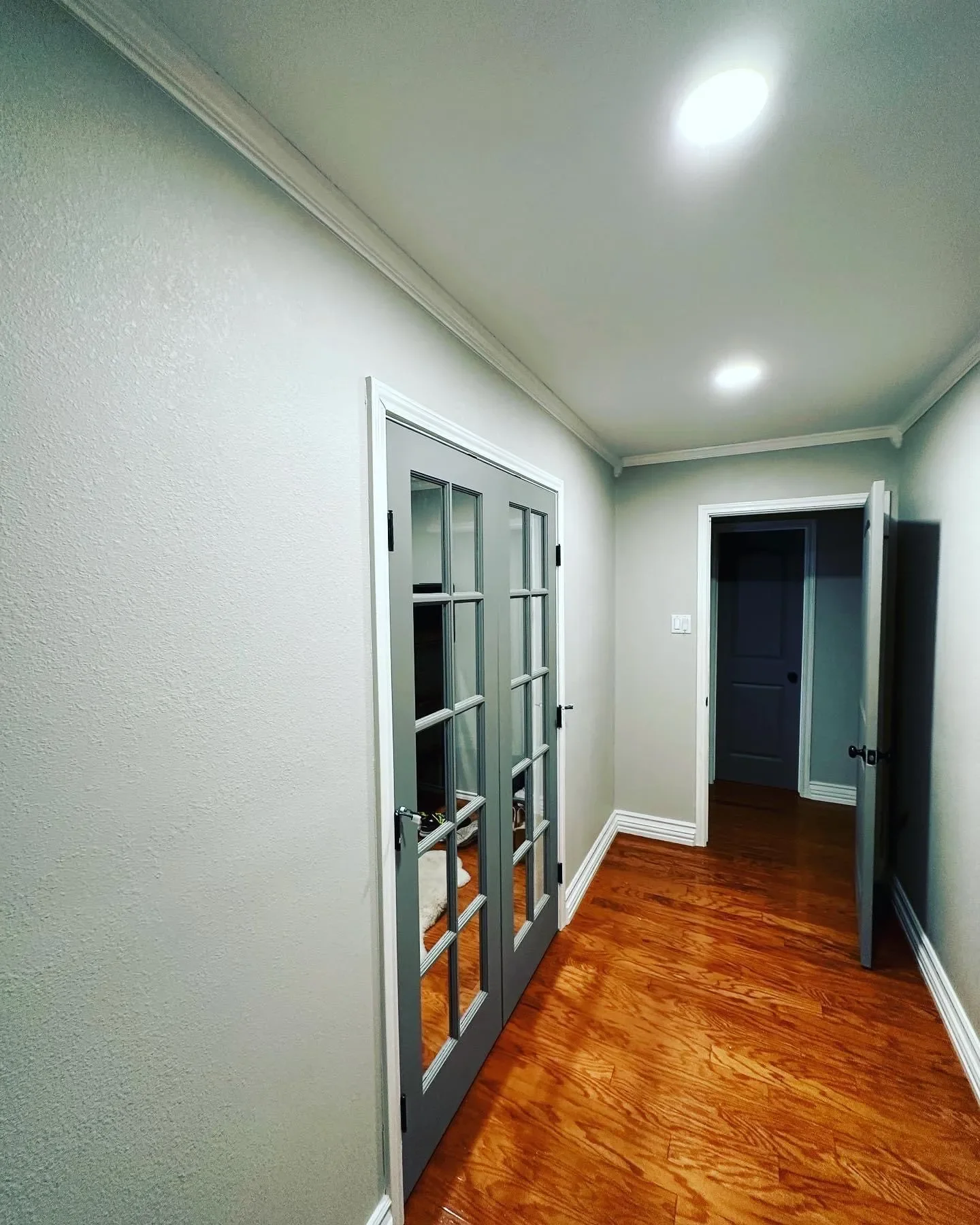 Empty hallway with white textured walls, hardwood flooring, two ceiling lights, a glass-paneled French door on the left, and an open door at the end of the hall leading to a dark room.
