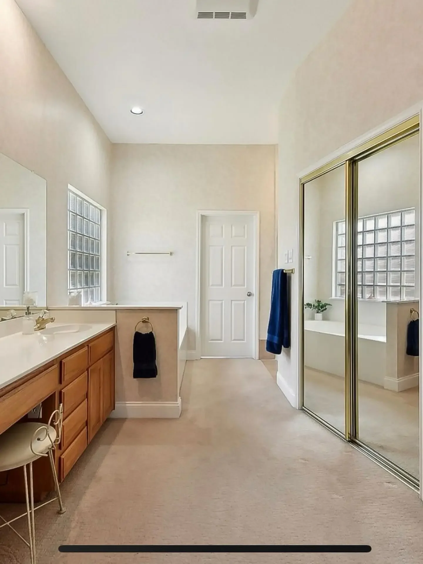A spacious bathroom with a double sink vanity, a large mirror, a glass block window, a closet with mirrored sliding doors, and a bathtub near a window with a small plant on the ledge.