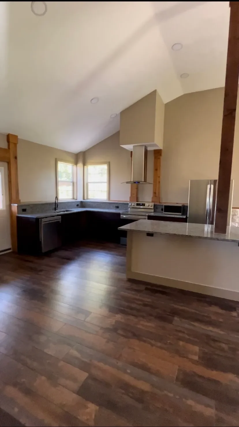 A modern kitchen with hardwood floors, beige walls, a dark lower cabinet set with a granite countertop, and a kitchen island. Two windows provide natural light, and there is a stainless steel stove with a vent hood above.