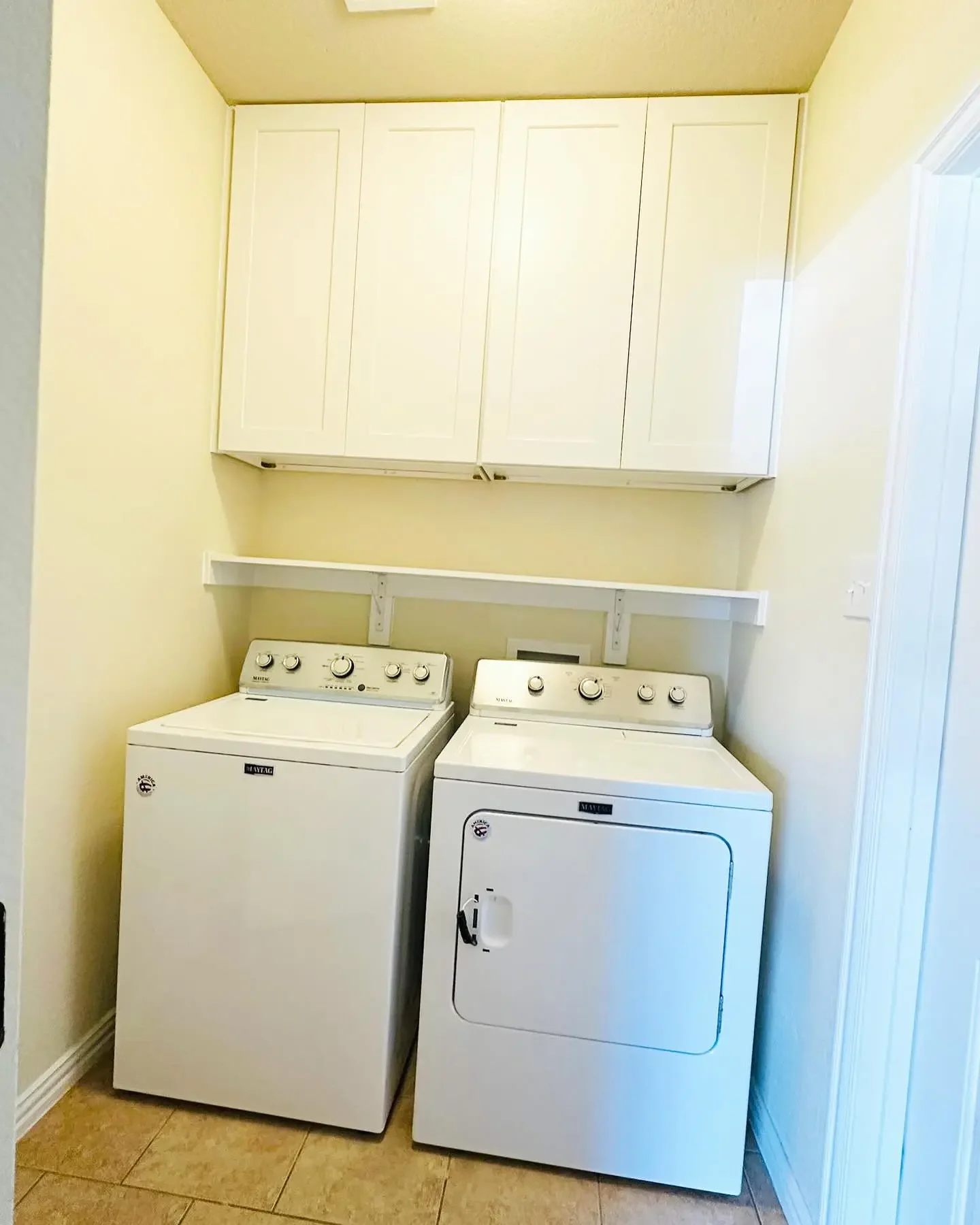A laundry room with a white top-loading washing machine and a white front-loading dryer, beige tiled floor, set against cream-colored walls with white cabinets above the appliances.