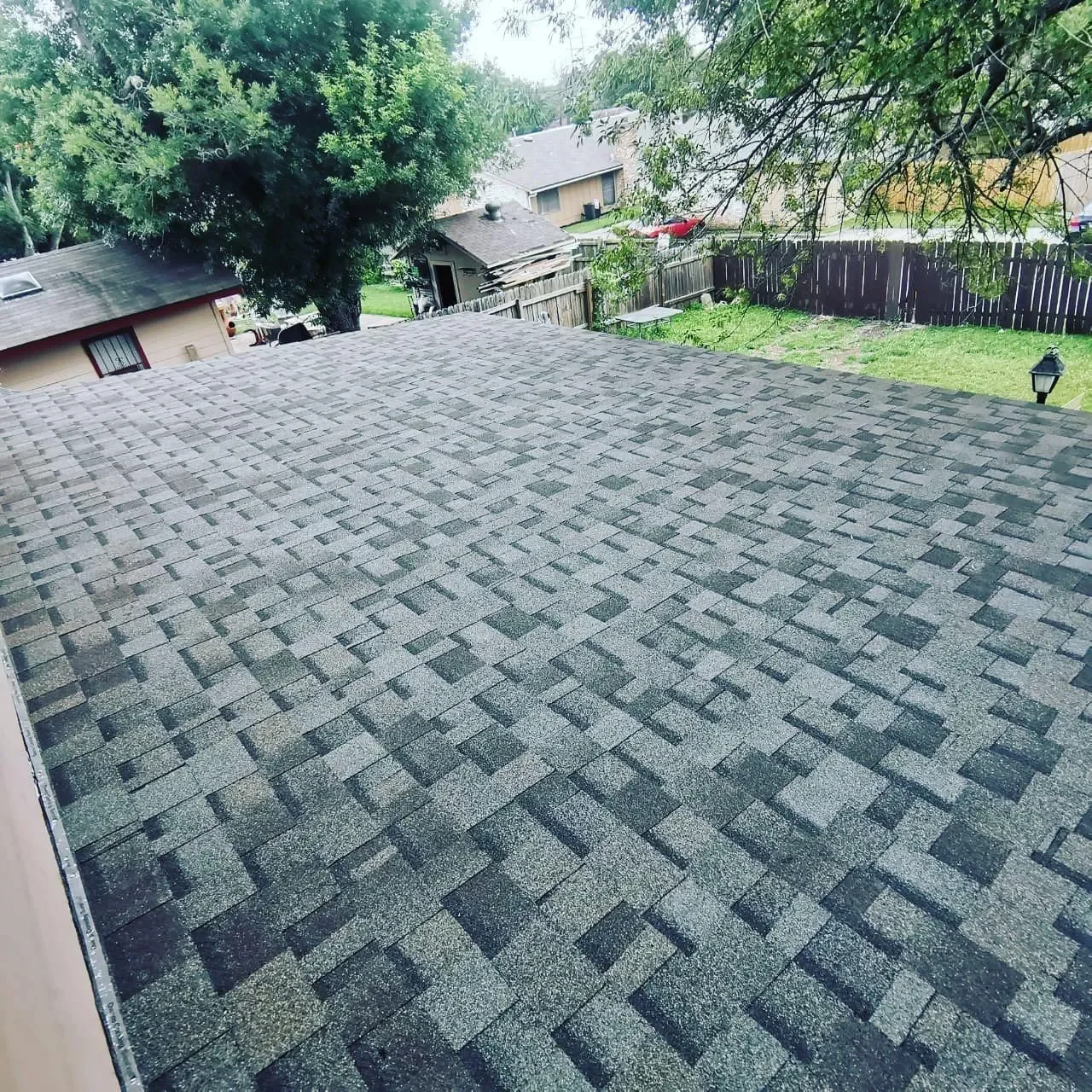 View of a rooftop with asphalt shingles, overlooking a backyard with grass, a wooden fence, trees, and neighboring houses.