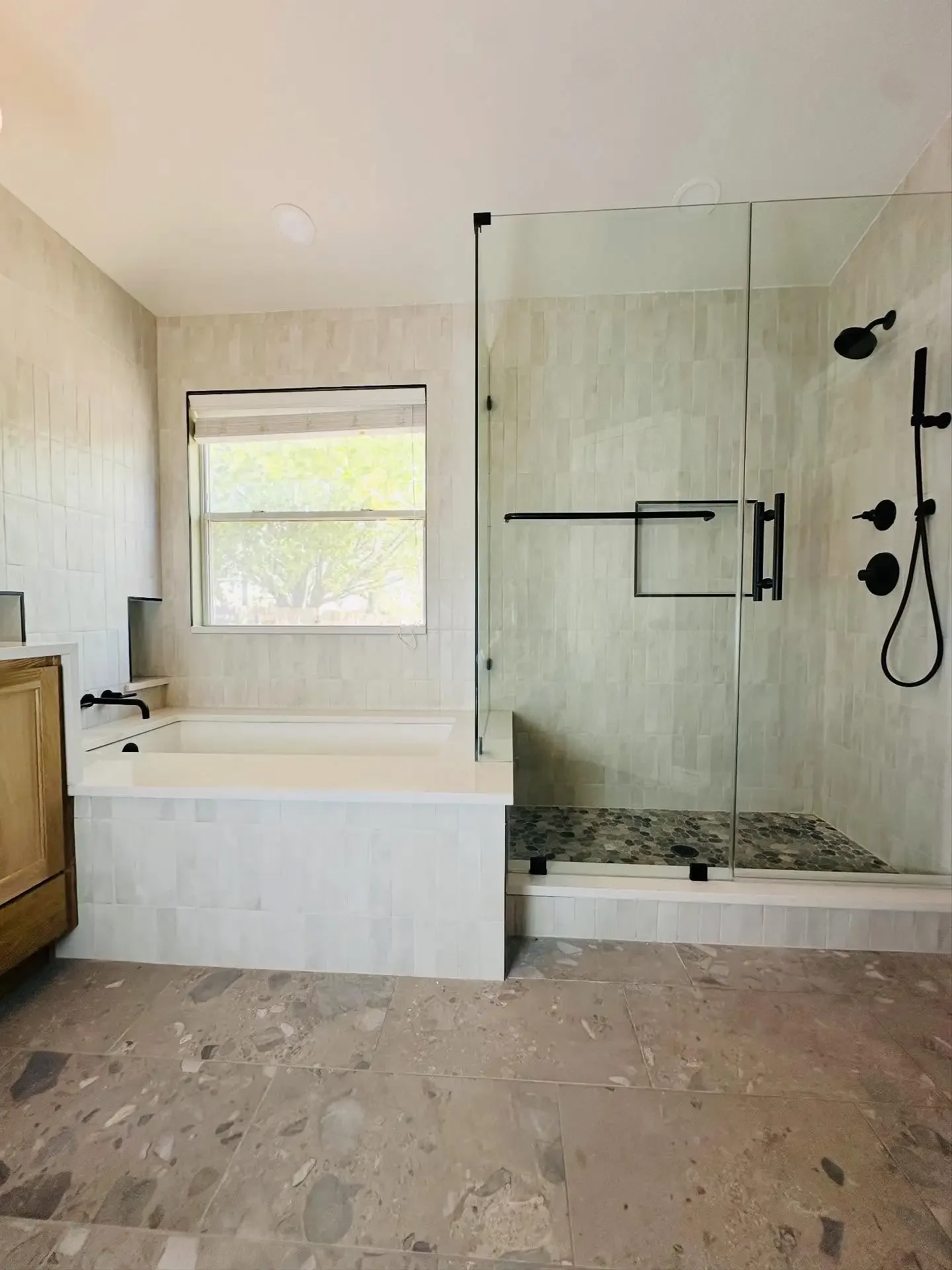 Modern bathroom featuring a built-in bathtub, a glass-enclosed shower with black fixtures, a window above the bathtub, beige tiled walls, and a stone tile floor.