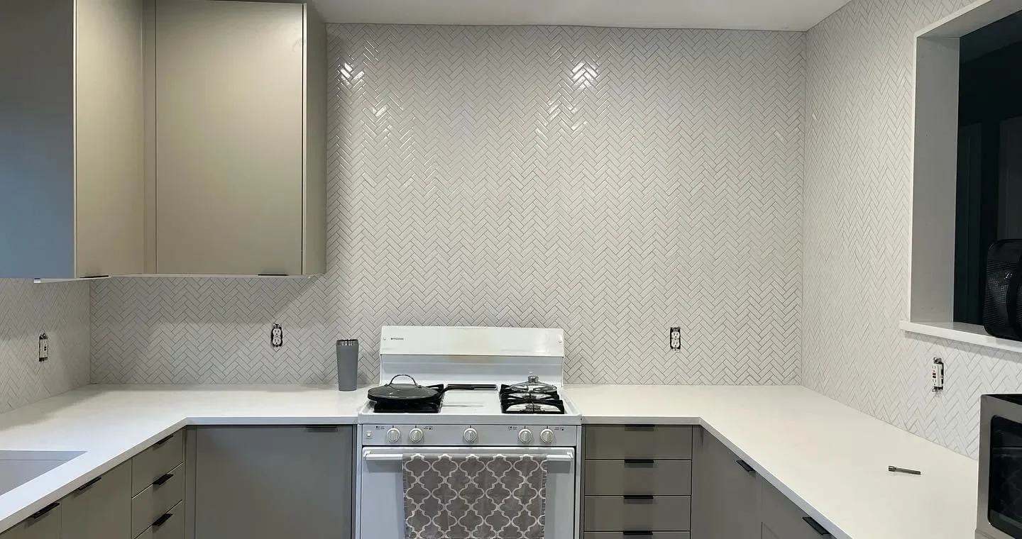Kitchen with gray cabinets, white countertops, and a gray and white patterned backsplash. A stove with a pot and a black travel mug on the counter, and a pen on the right side of the counter.