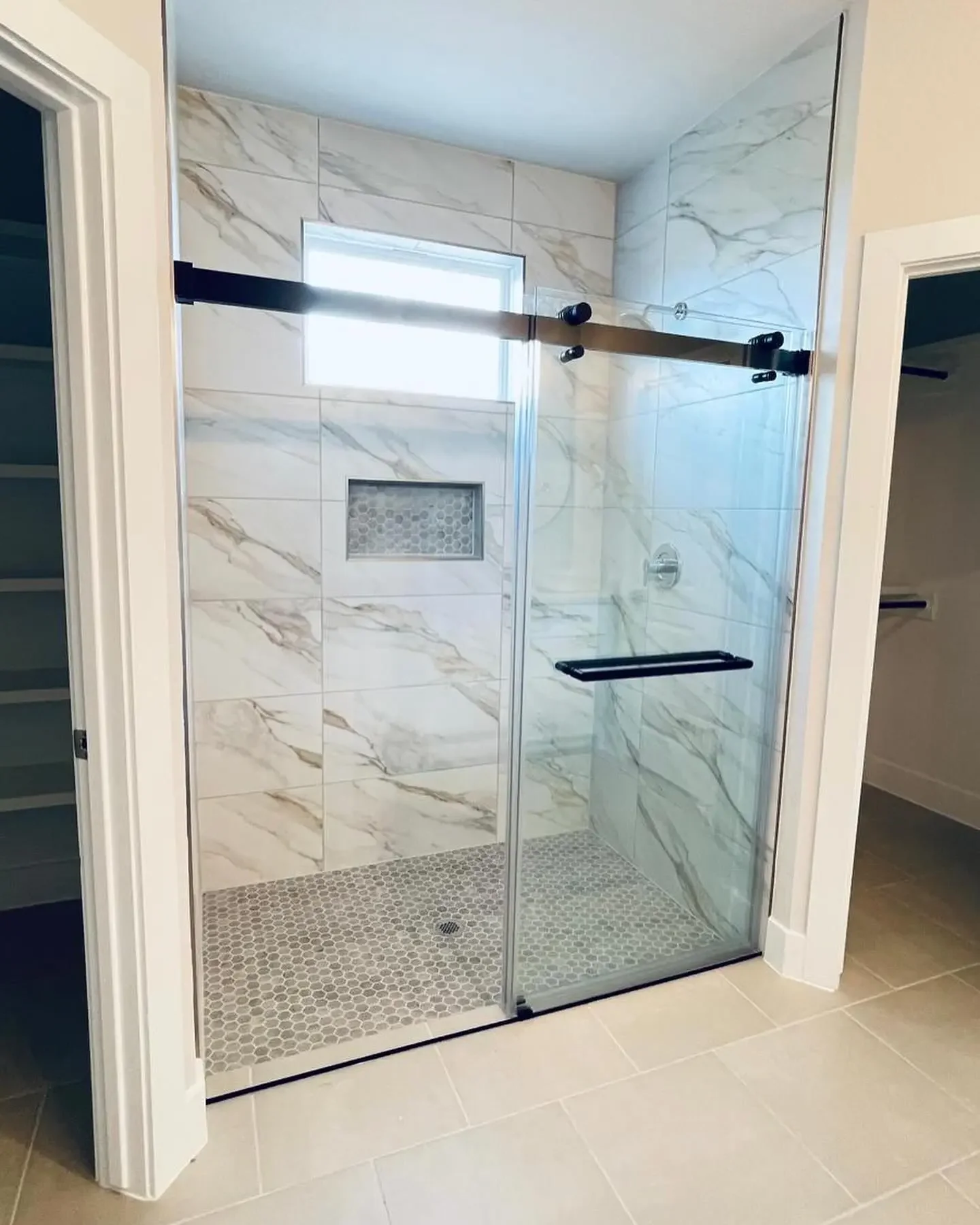 Modern glass shower with marble tiles, a built-in niche, and a small window; has a black handle and sliding door, with beige tile flooring outside.