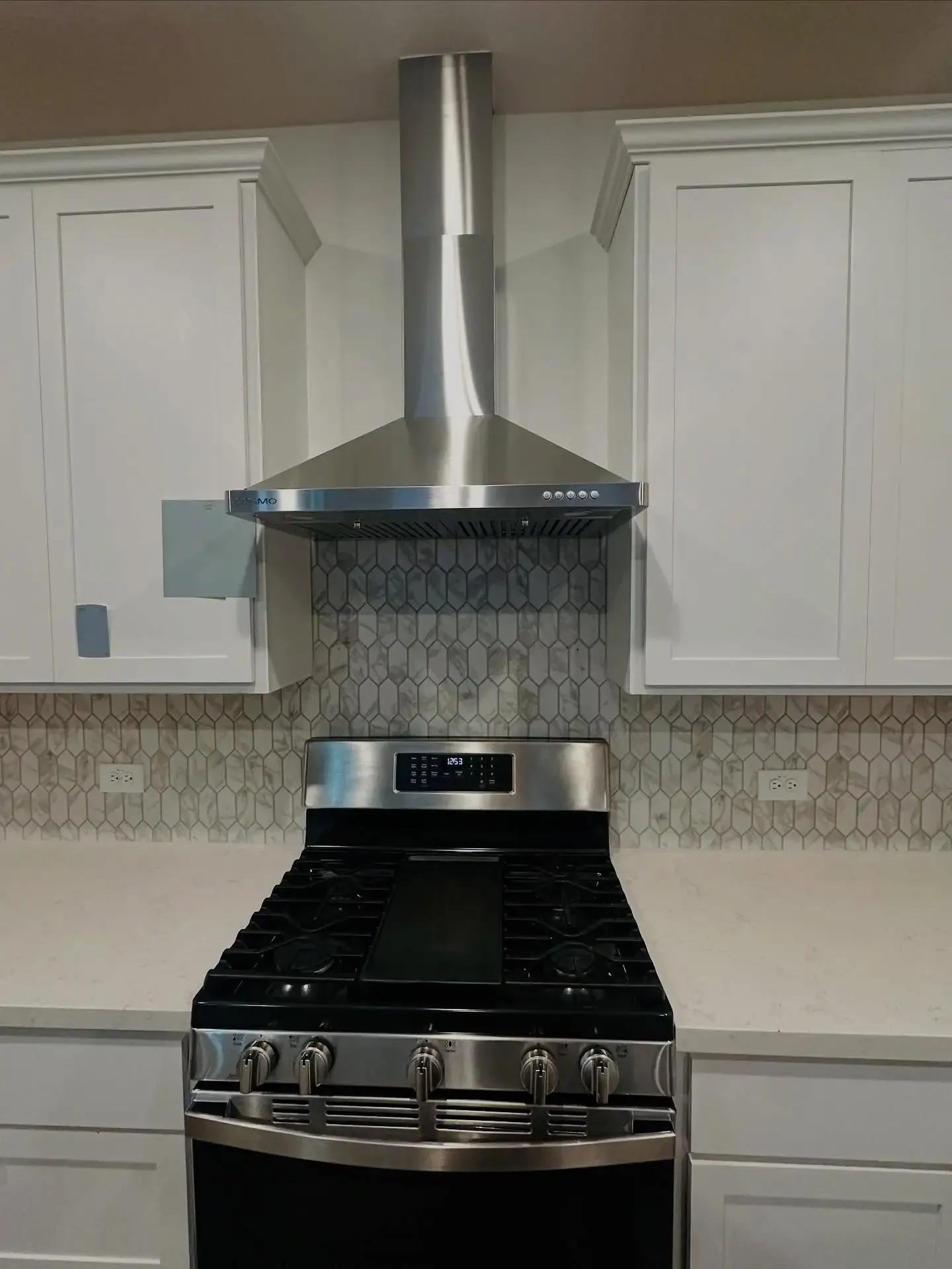 Stainless steel range with black cooktop and control knobs, mounted under a modern stainless steel range hood with a hexagon tile backsplash in a kitchen with white cabinets.
