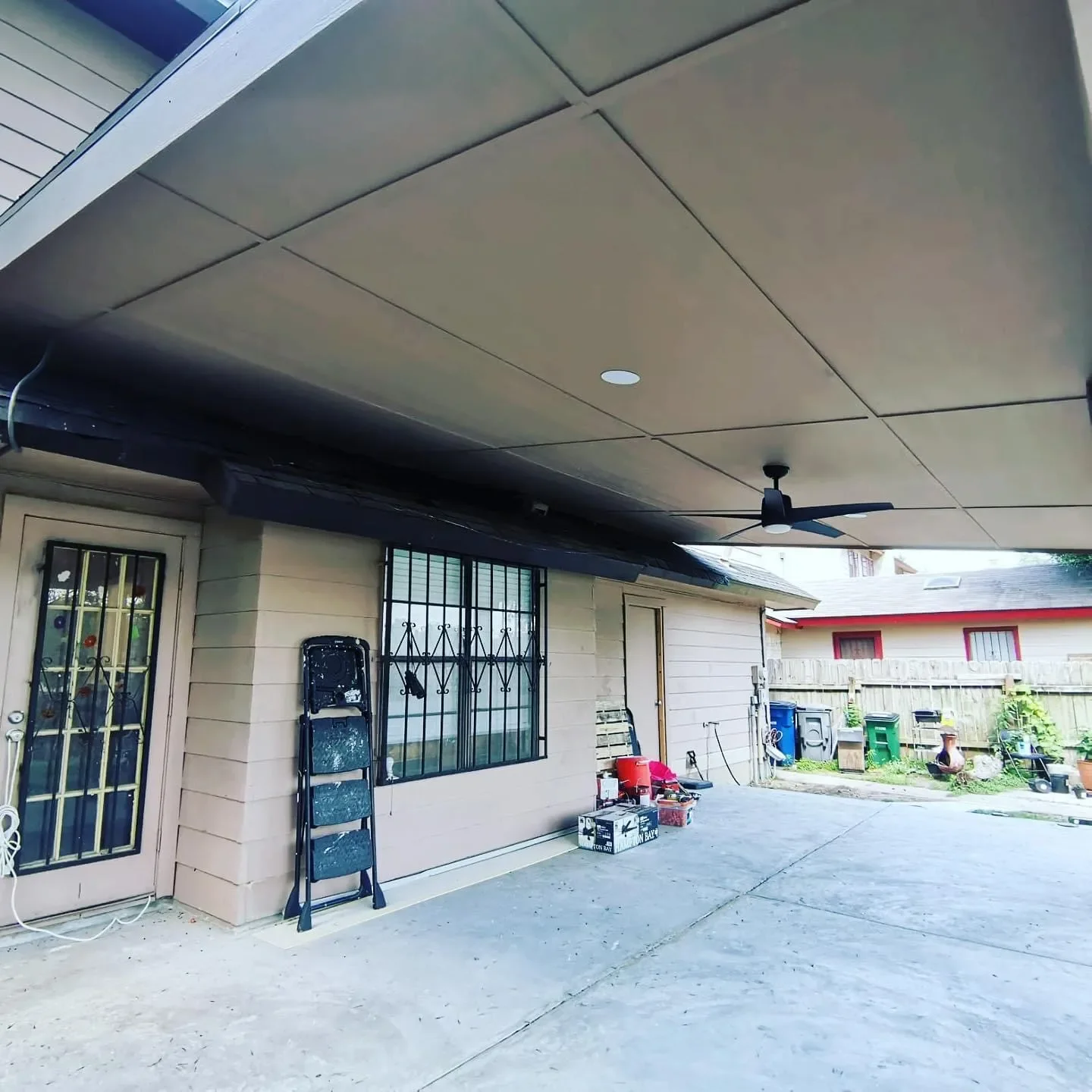 Backyard patio area with a beige house wall, a black ceiling fan, and various items like a folding ladder, boxes, and outdoors equipment. A fenced backyard with some plants and garden decorations is visible in the background.