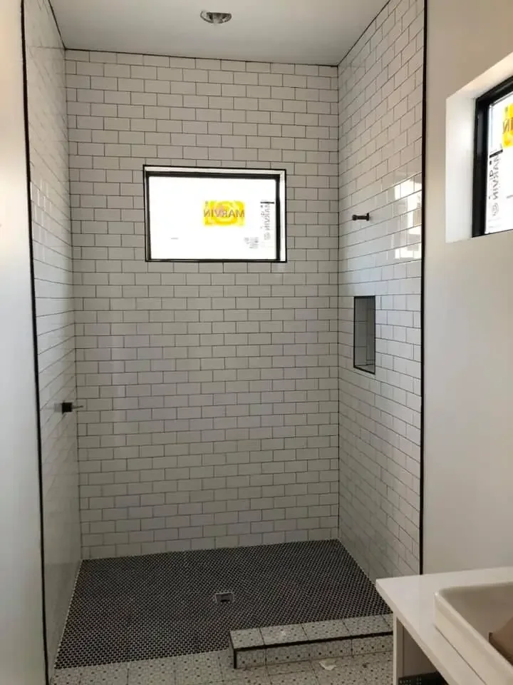 A partially constructed walk-in shower with white subway tile walls and small black mosaic tile floor. There is a window on the back wall and an opening for a shower niche.