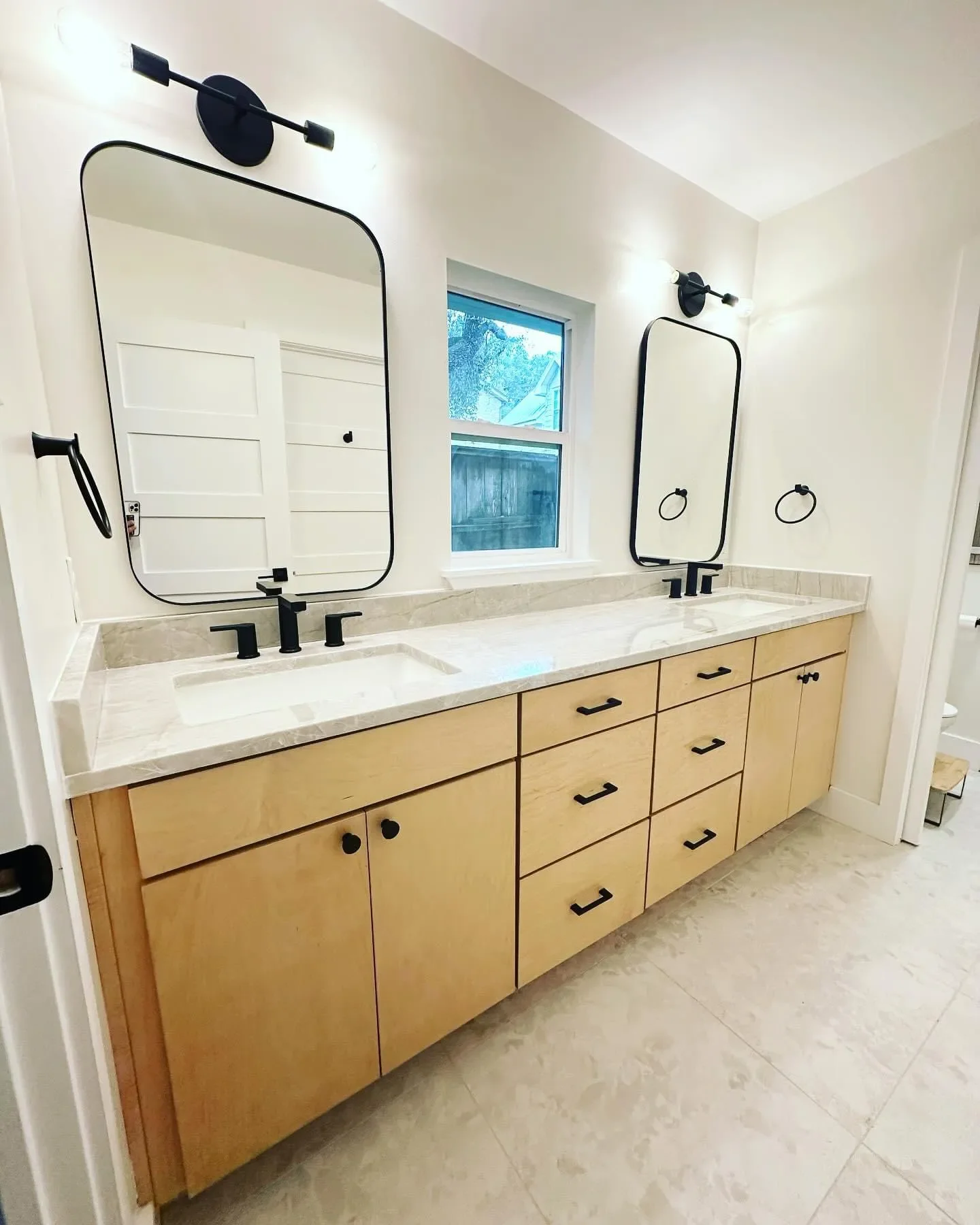 A modern bathroom vanity with double sinks, black fixtures, and large rectangular mirrors. The vanity has light wood cabinets with black handles, and a marble countertop. Two black light fixtures and two black towel rings are mounted on the white wal