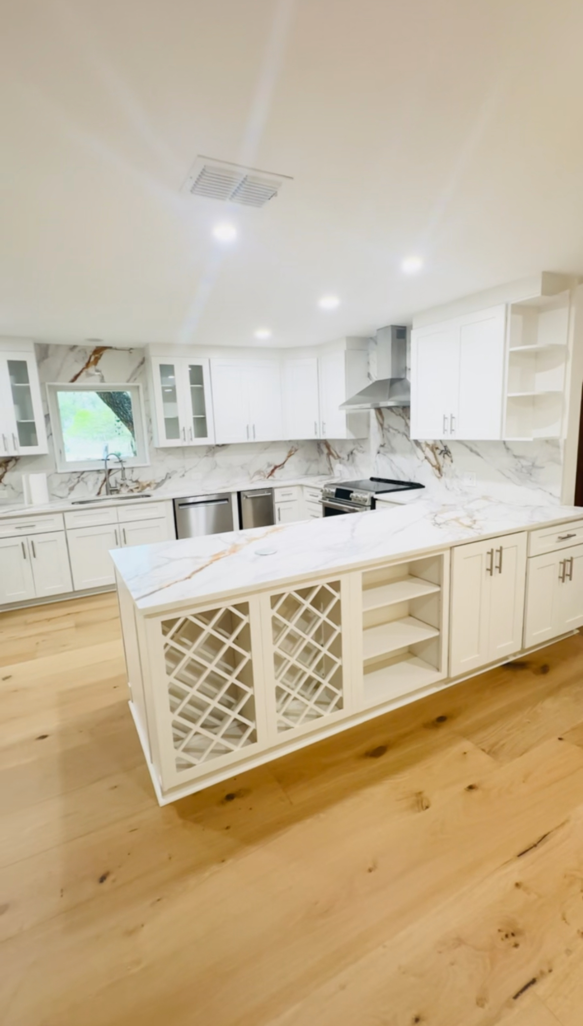 A modern white kitchen with marble countertops, white cabinets, and a wood floor, featuring a kitchen island with wine rack storage and open shelves.