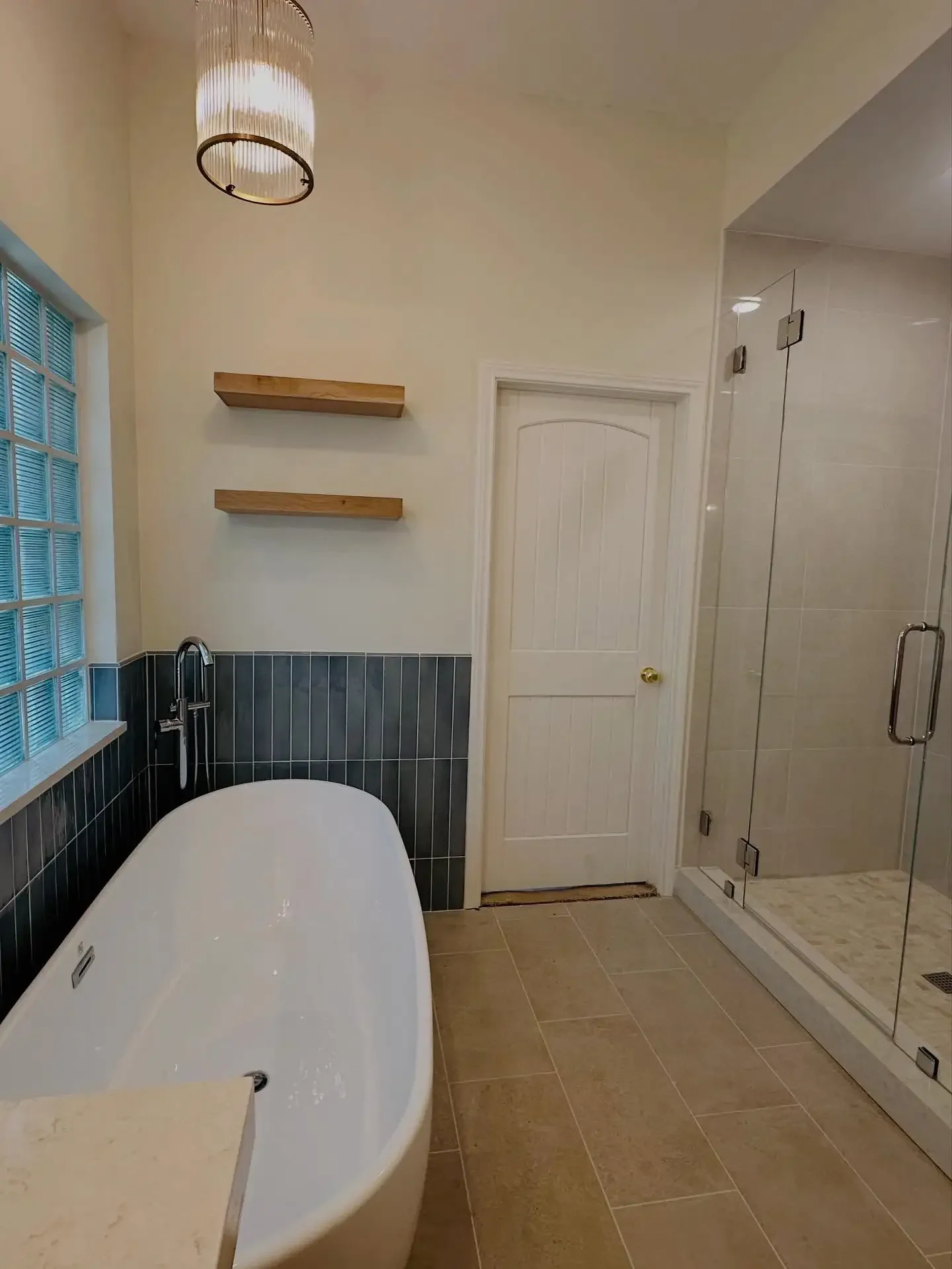 A modern bathroom with a freestanding bathtub, a glass-enclosed shower, a door, tiled walls, and a window with glass blocks. Two wooden shelves are mounted on the wall above the bathtub, and a ceiling light fixture is visible.