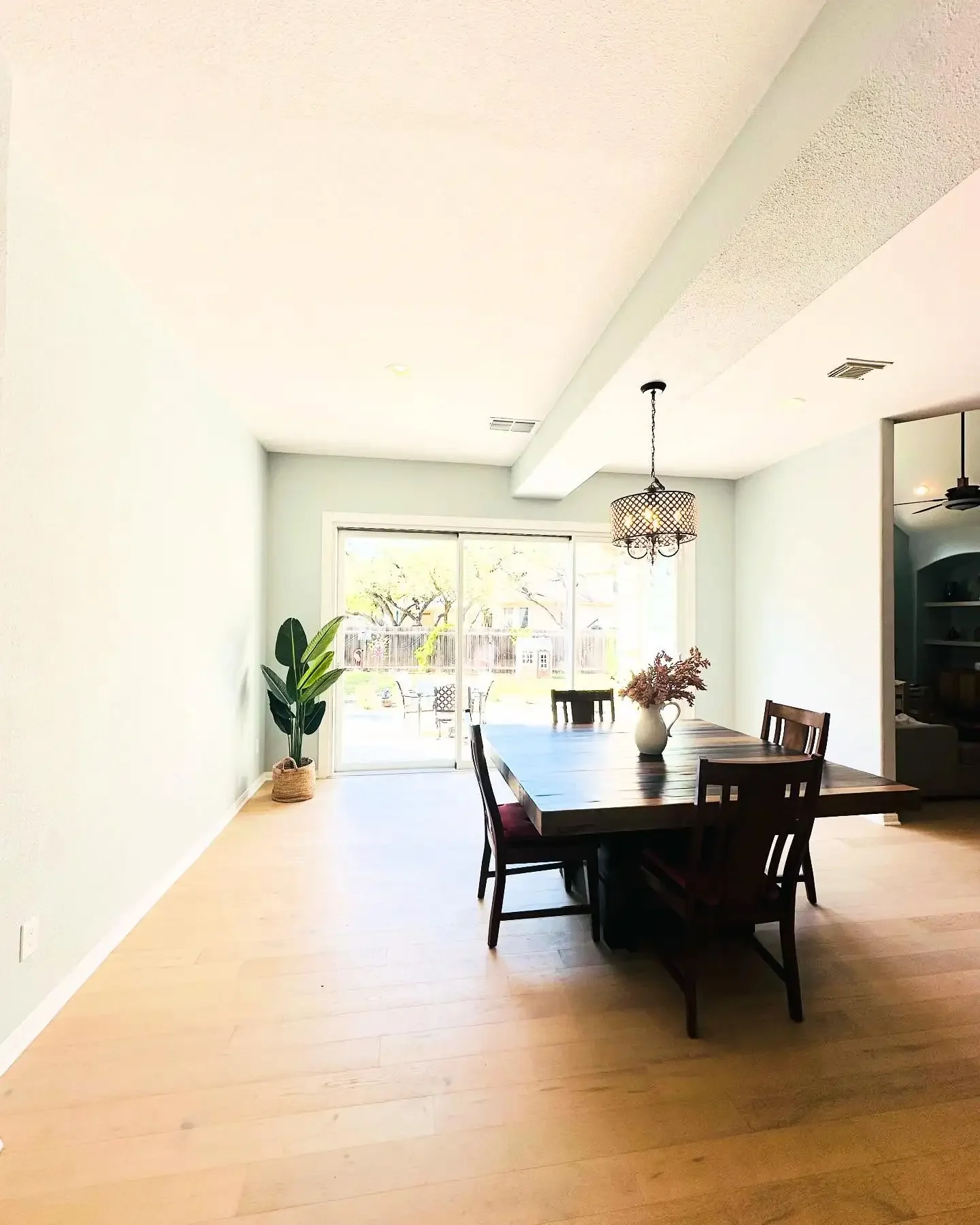 A dining room with a large wooden table, four chairs, a hanging chandelier, a sliding glass door leading to a backyard, a potted plant in the corner, and sunlight coming through the windows.