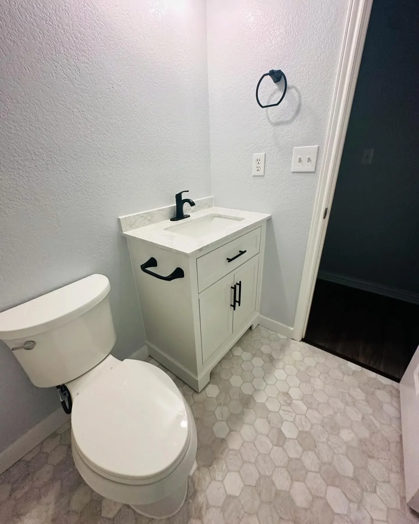 Small bathroom with a white toilet and a white vanity with a black faucet, white hexagonal tile flooring, and a black towel ring on a white wall.