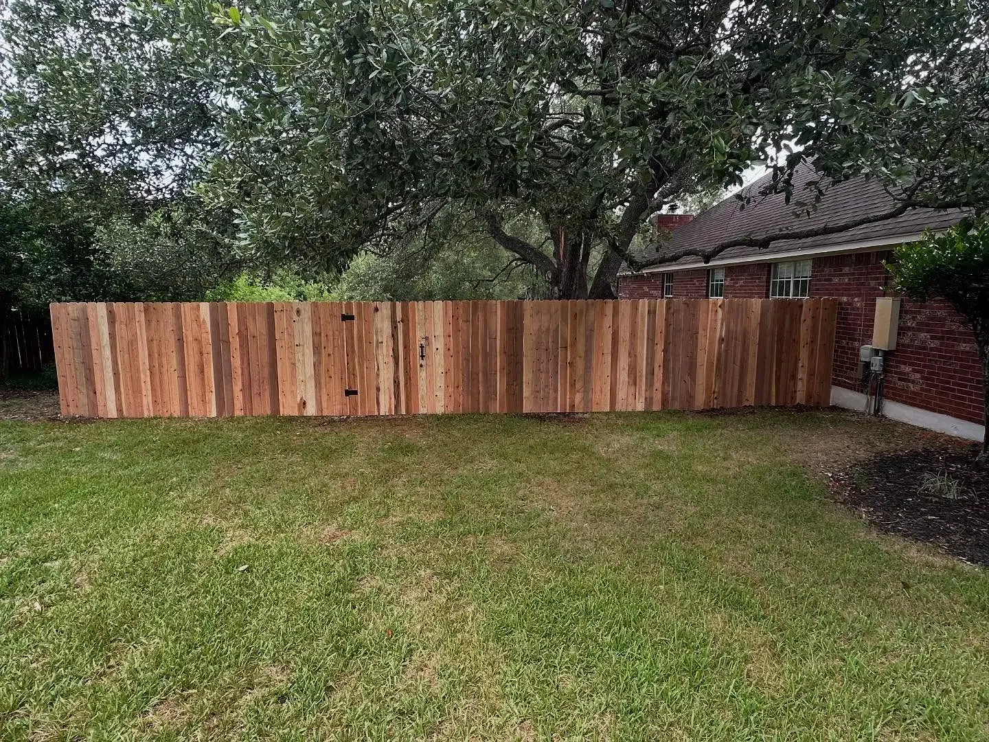 New wooden privacy fence installed in a backyard, with a brick house and trees in the background.