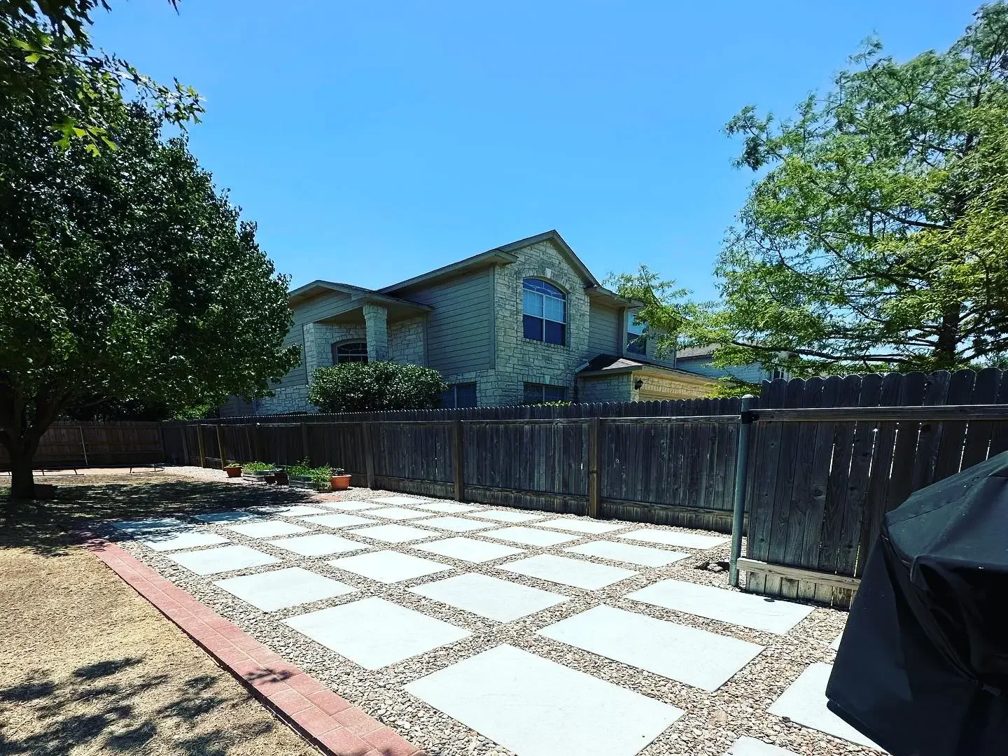 Backyard with stone pathway, wooden fence, trees, house in the background, and a covered grill in the corner.