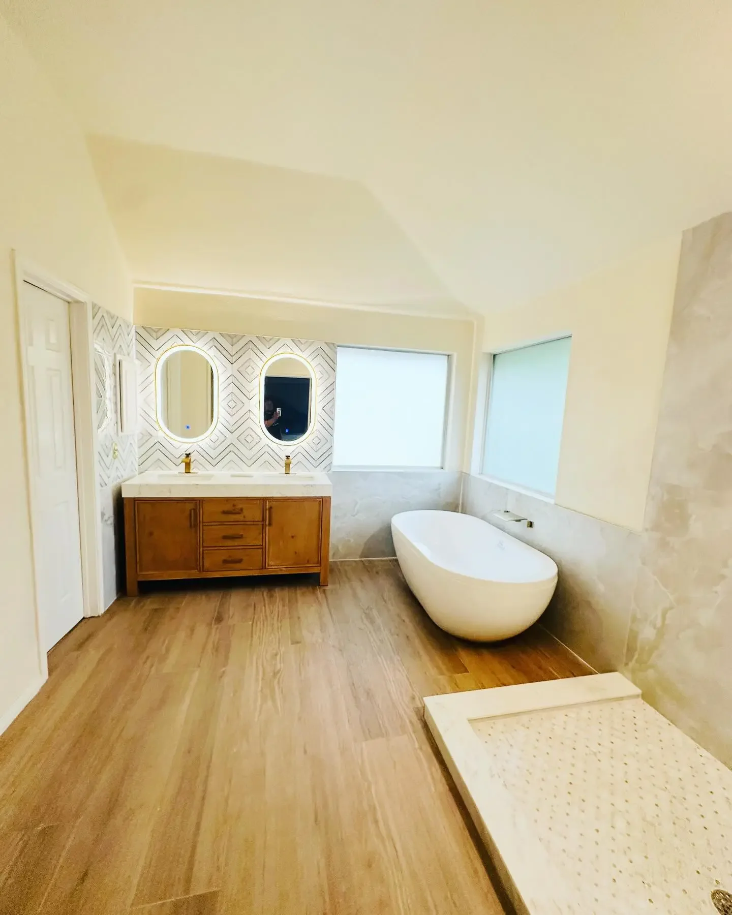 Modern bathroom with wooden vanity, two oval mirrors, a freestanding bathtub, and a shower tray, featuring light colors and wood flooring.