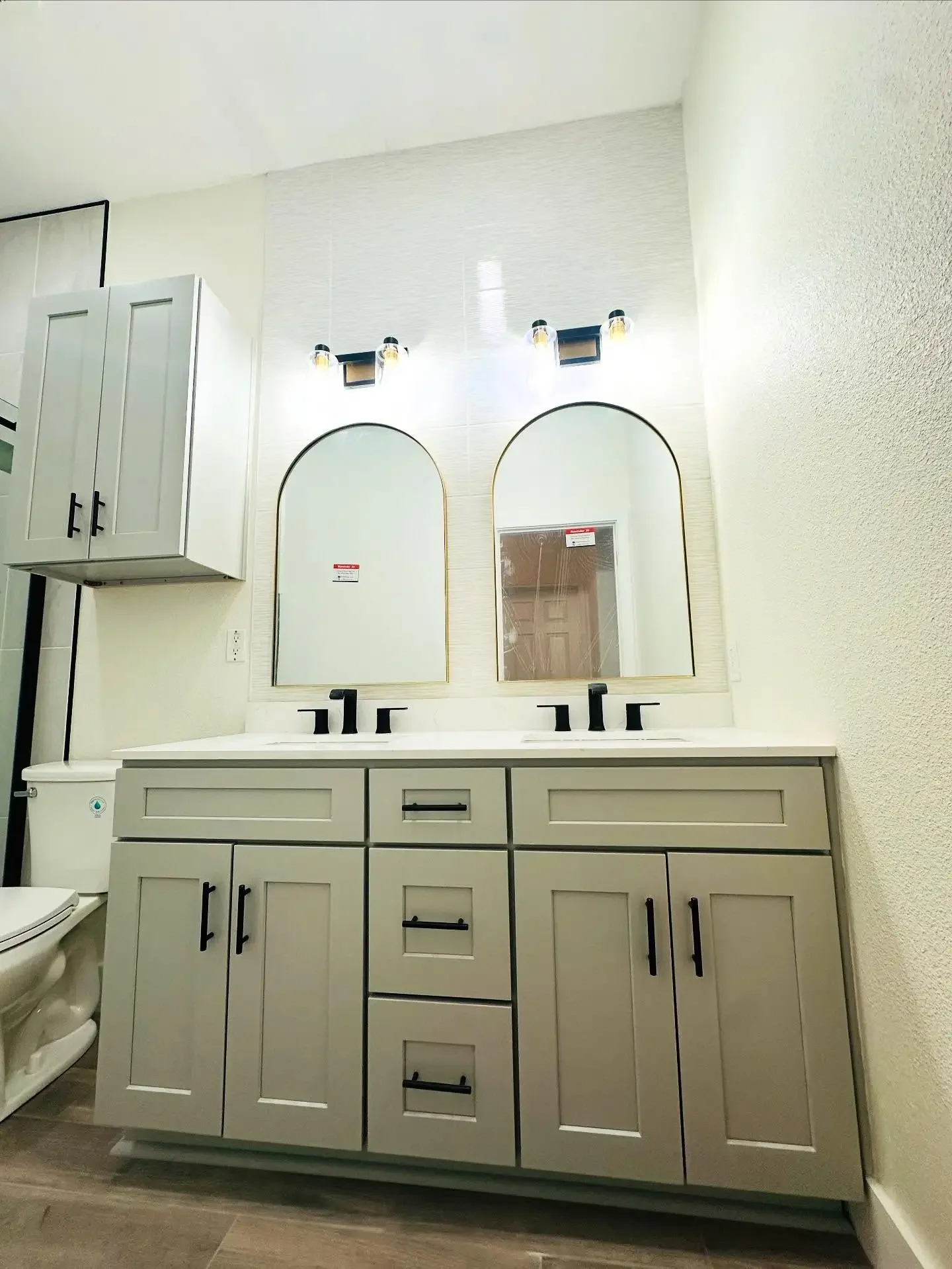 Double bathroom vanity with two black faucets and two large mirrors with rounded tops, above a light gray cabinet with black handles, in a bathroom with white textured walls.