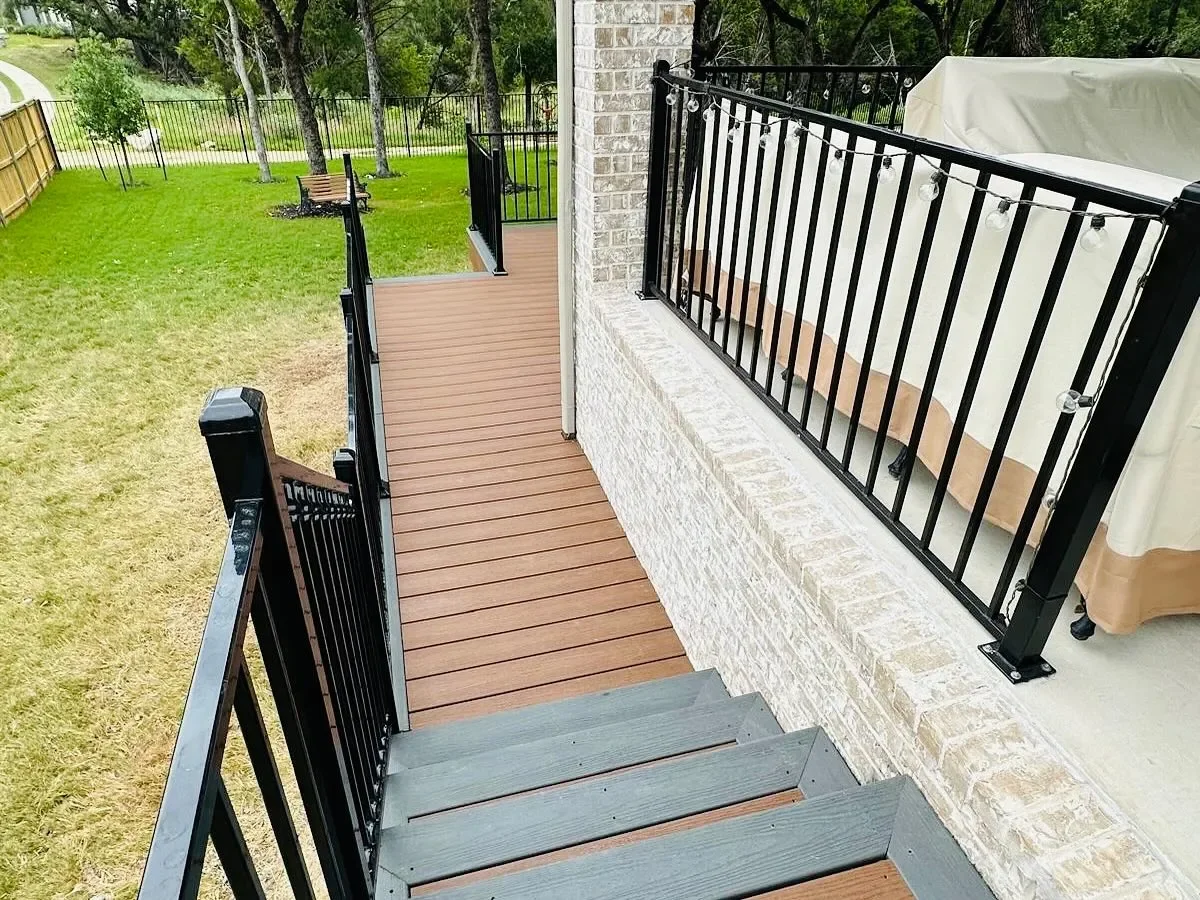 View of a backyard with a deck and stairs, black metal railing, green grass, and trees in the background.