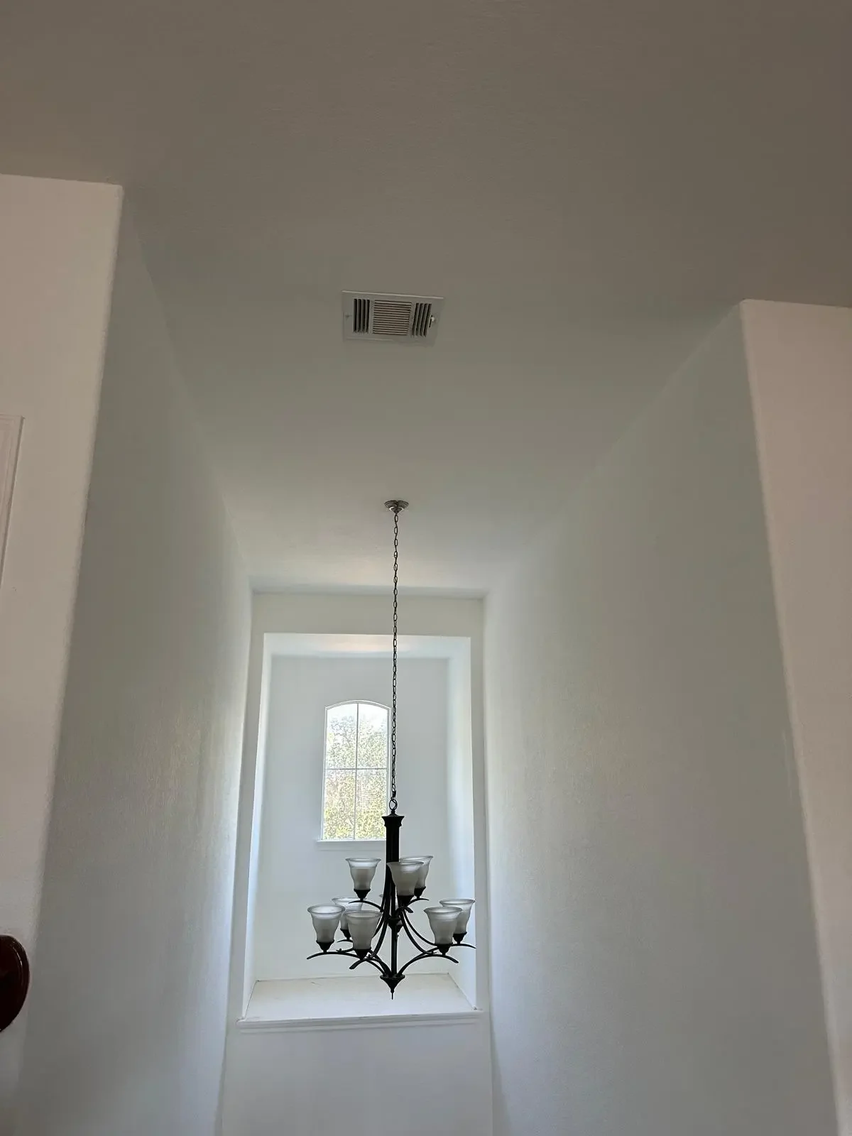 Interior view of a house hallway with a chandelier hanging from the ceiling and a window at the end letting in natural light.