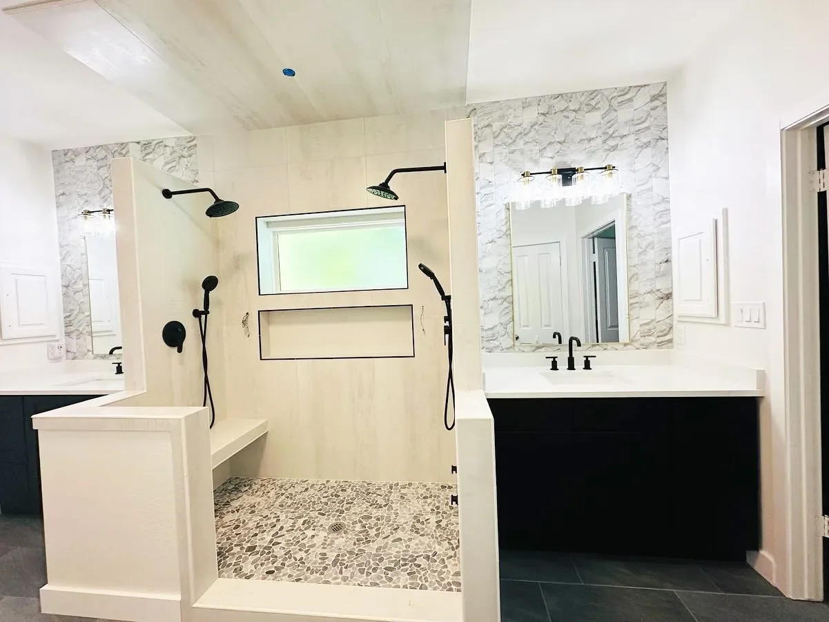 Modern bathroom with a walk-in shower featuring black fixtures, a pebble tile floor, and a large mirror above a white vanity with black cabinetry.