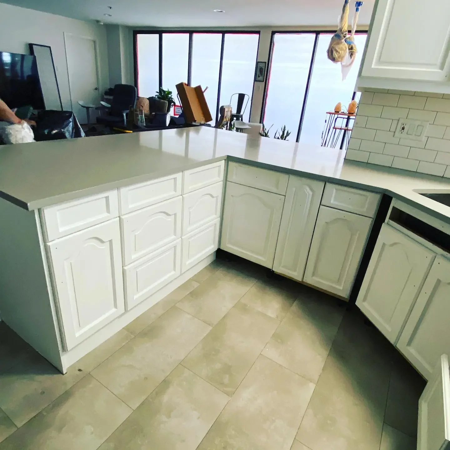 White kitchen cabinets with beige countertops, tiled floor, and a large window in the background.