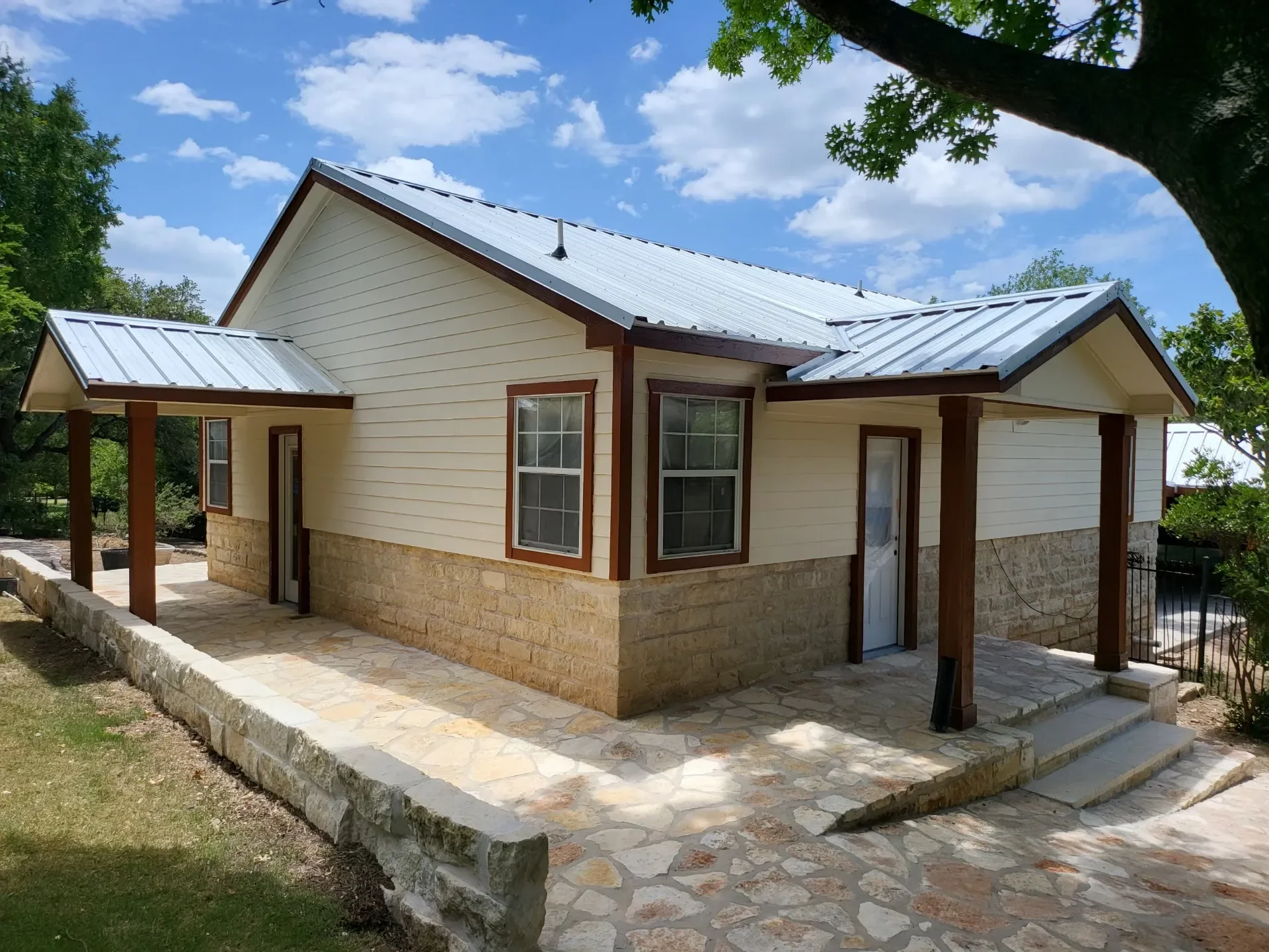 A small house with a metal roof, cream-colored siding, stone foundation, wooden columns, and a stone paved porch.