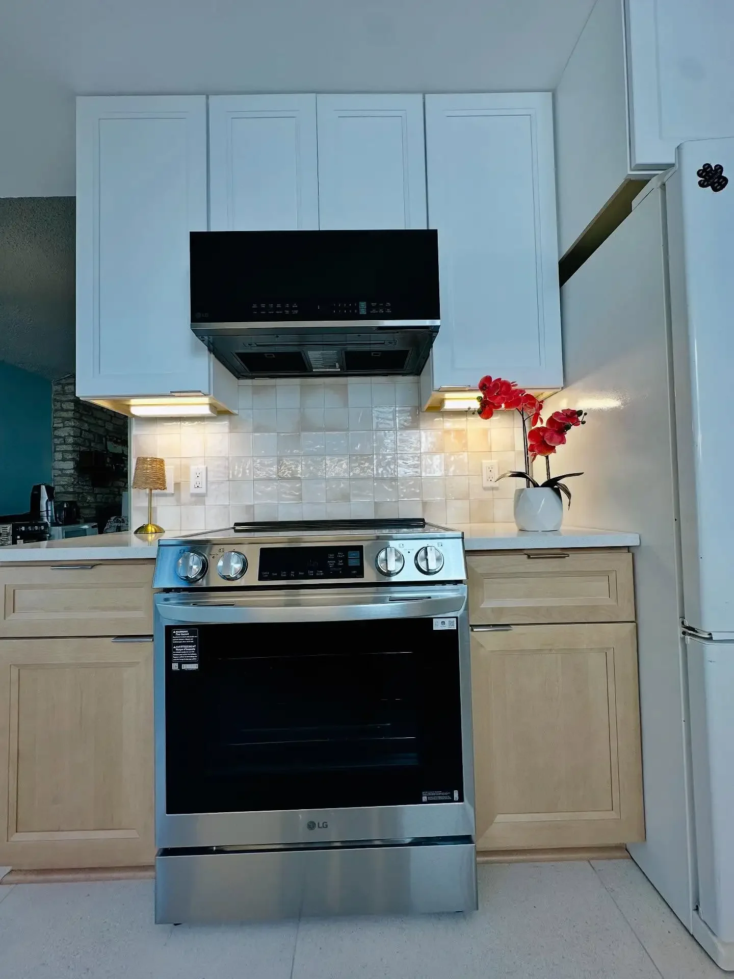 Kitchen with stainless steel oven, white cabinets above, white tiled backsplash, white countertop with a vase of red flowers, and part of a white refrigerator to the right.