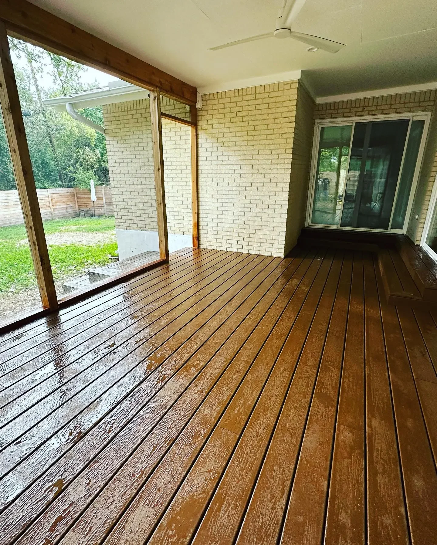 Covered porch with wet wooden decking, brick walls, sliding glass door, ceiling fan, and ceiling.