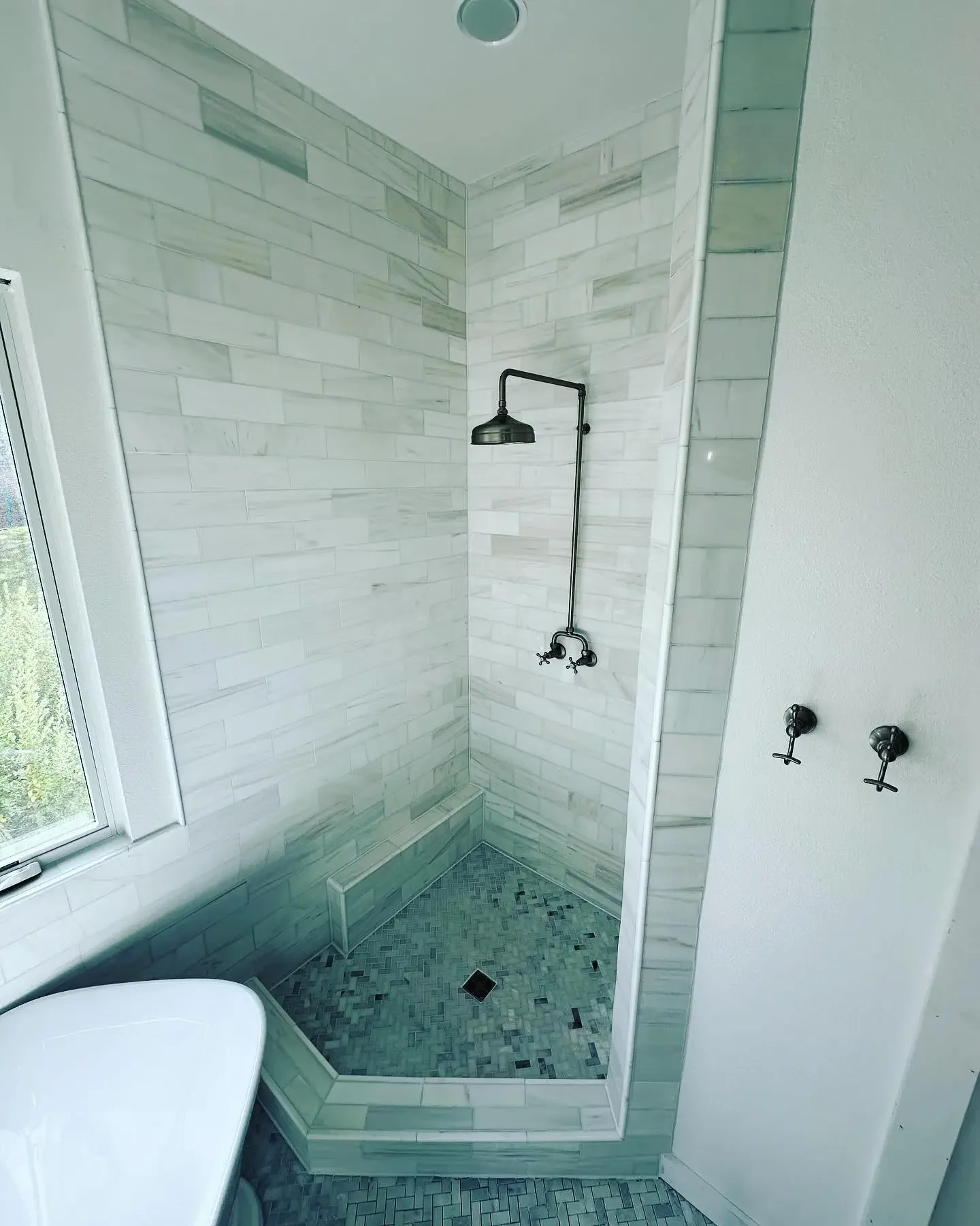 Shower area with white subway tile walls, black showerhead, small mosaic floor tiles, a bench in the corner, a window on the left, and two hooks on the wall.