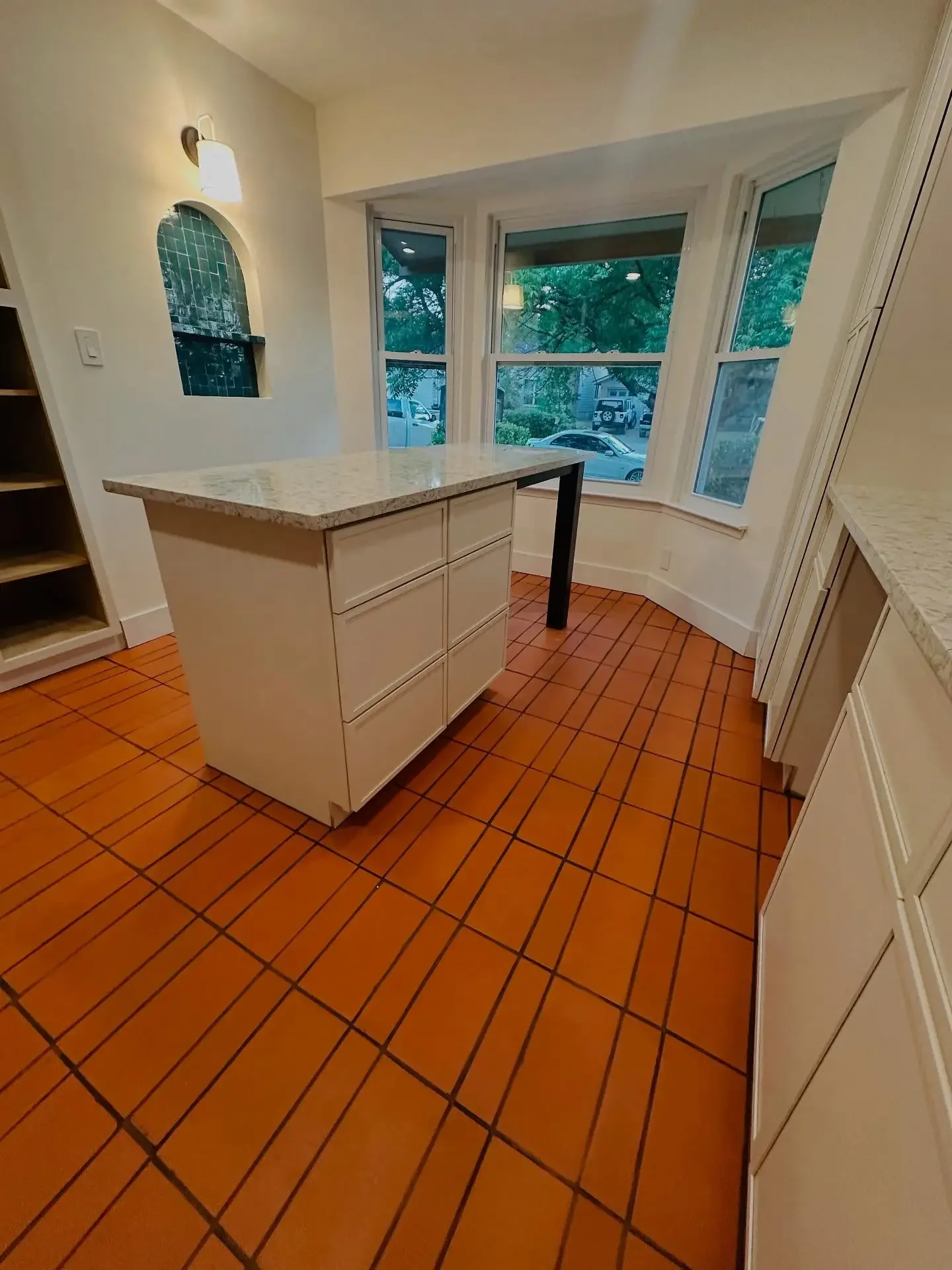 A kitchen with terracotta tile flooring, white cabinets, a granite countertop, large bay windows, and a small kitchen island with drawers, with a wall-mounted light fixture and an arched wall niche.