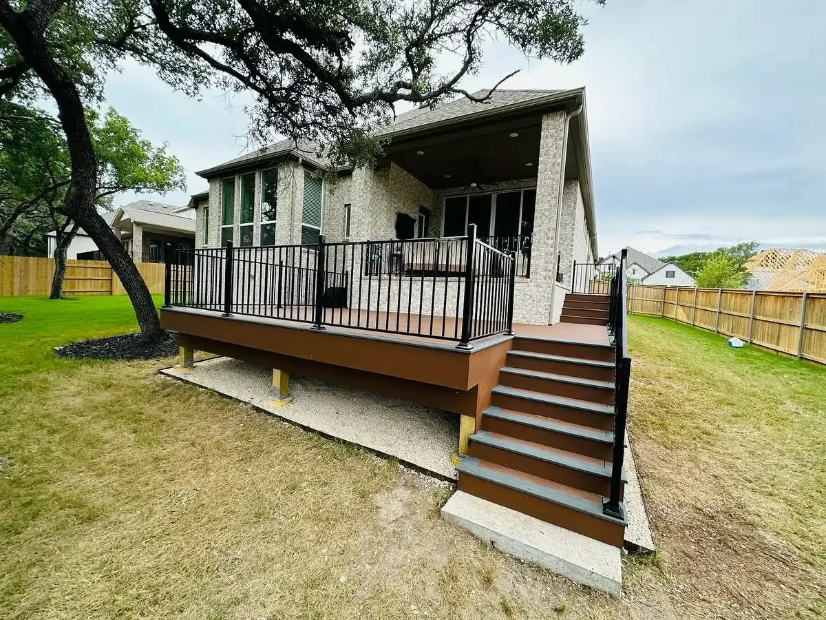 A new elevated wooden deck with black metal railings attached to the back of a house, with stairs leading down to the yard, in a backyard with a tree and a wooden privacy fence.