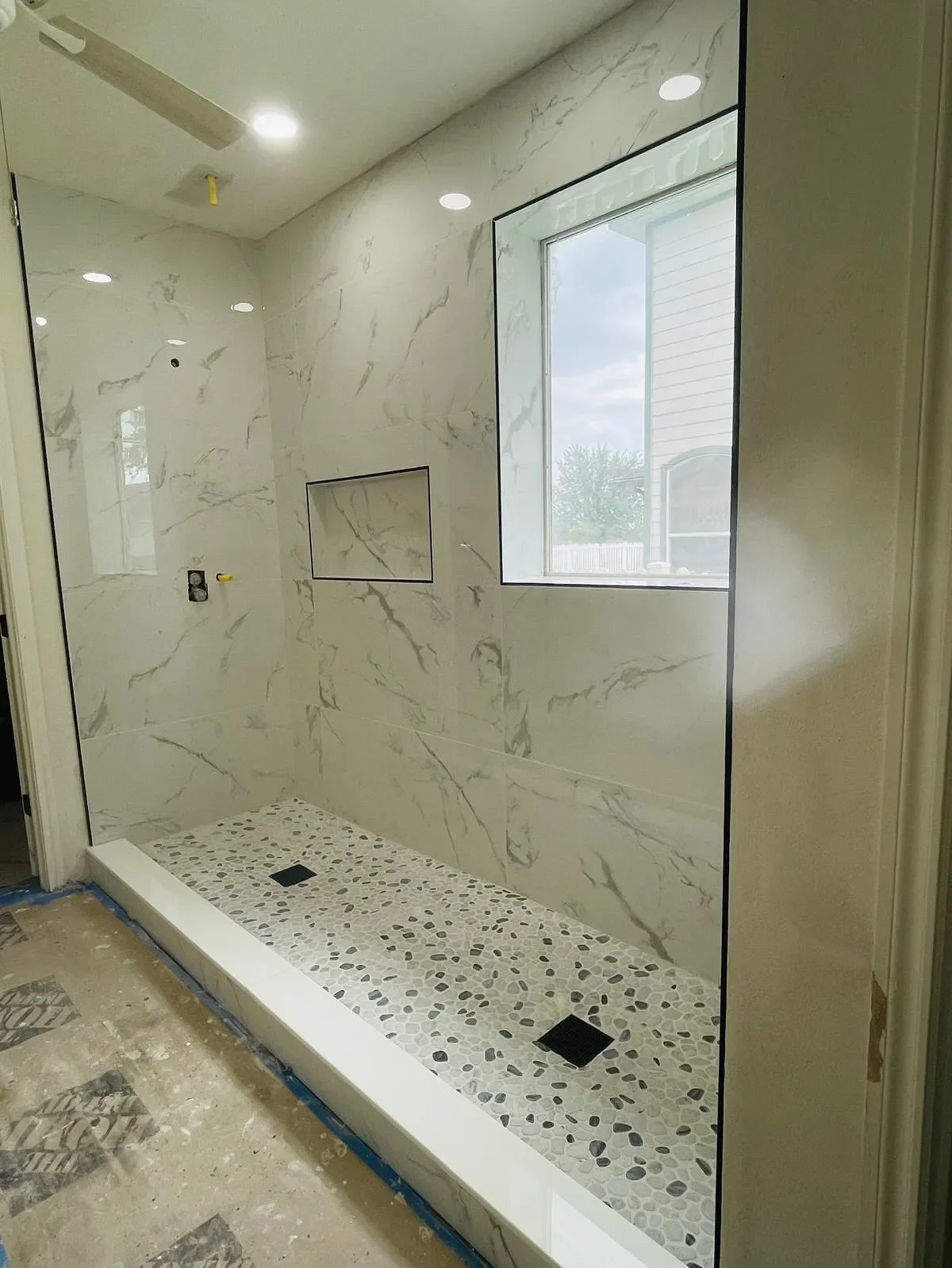 A modern walk-in shower with white marble walls, a pebble tile shower floor, a large window, and built-in niche for toiletries.