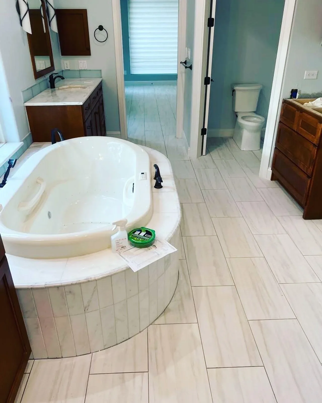 A bathroom with a large whirlpool bathtub, a wooden vanity with a sink, a toilet in a separate room, and a hallway leading to a door with blinds.